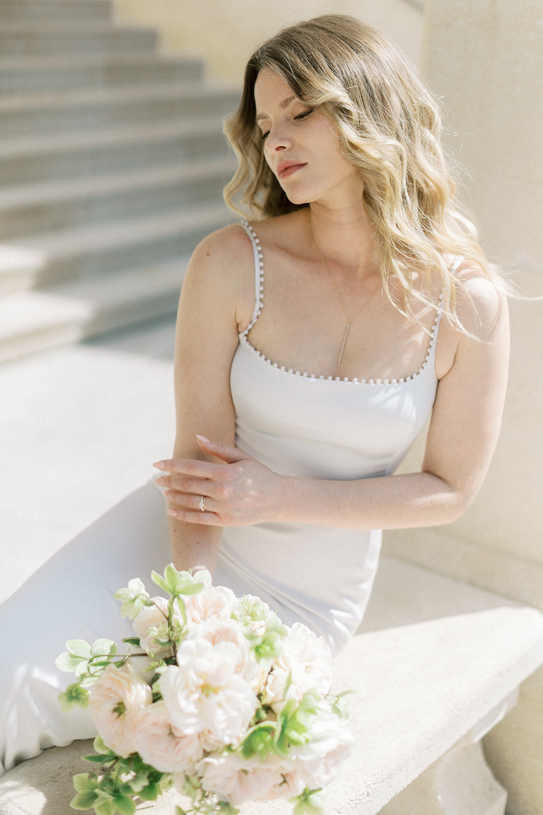 Bride seated outdoors in white spaghetti-strap slip dress with blush pink garden rose bouquet and diamond ring