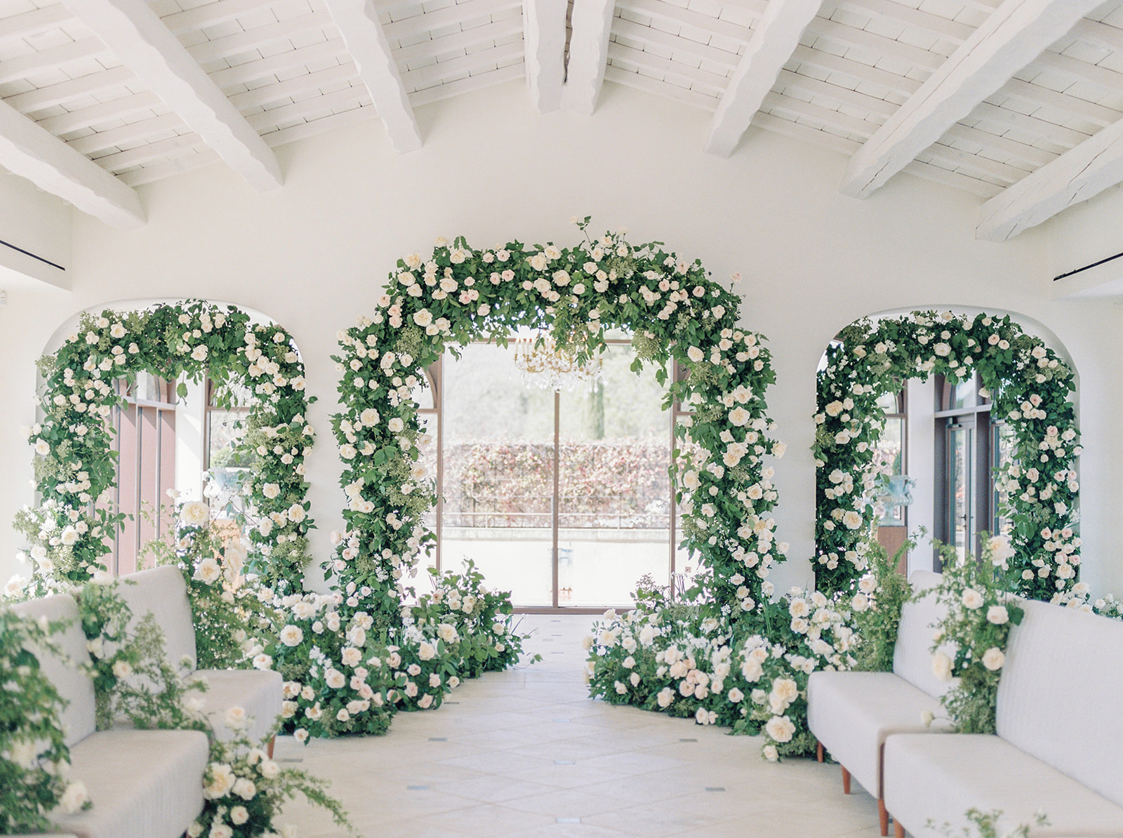 Indoor ceremony space with three floral-framed arches of ivory roses and white upholstered seating