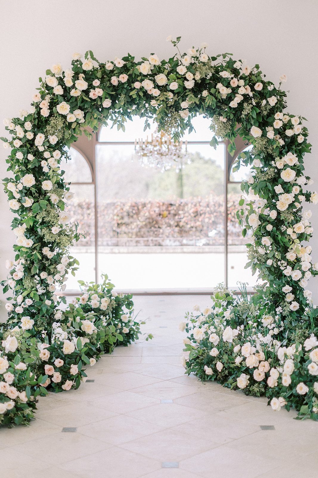 Circular floral arch of cream roses and white ranunculus installed before tall arched window indoors