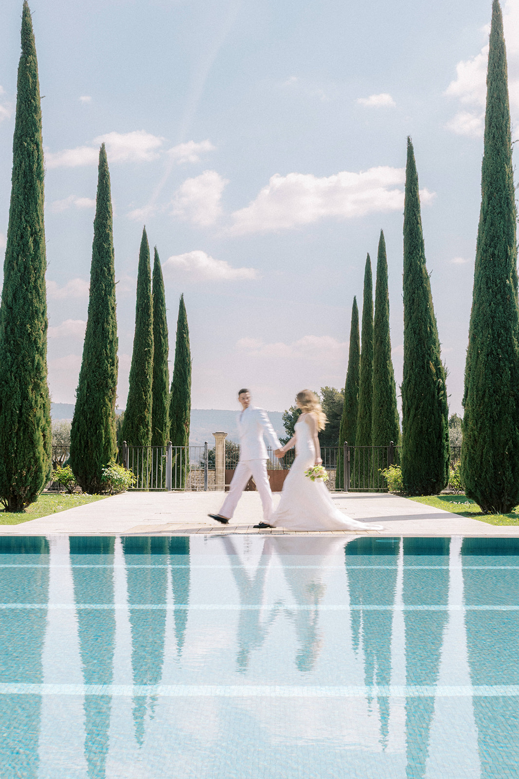 Bride and groom walking hand-in-hand beside infinity pool lined with cypress trees at Provencal estate