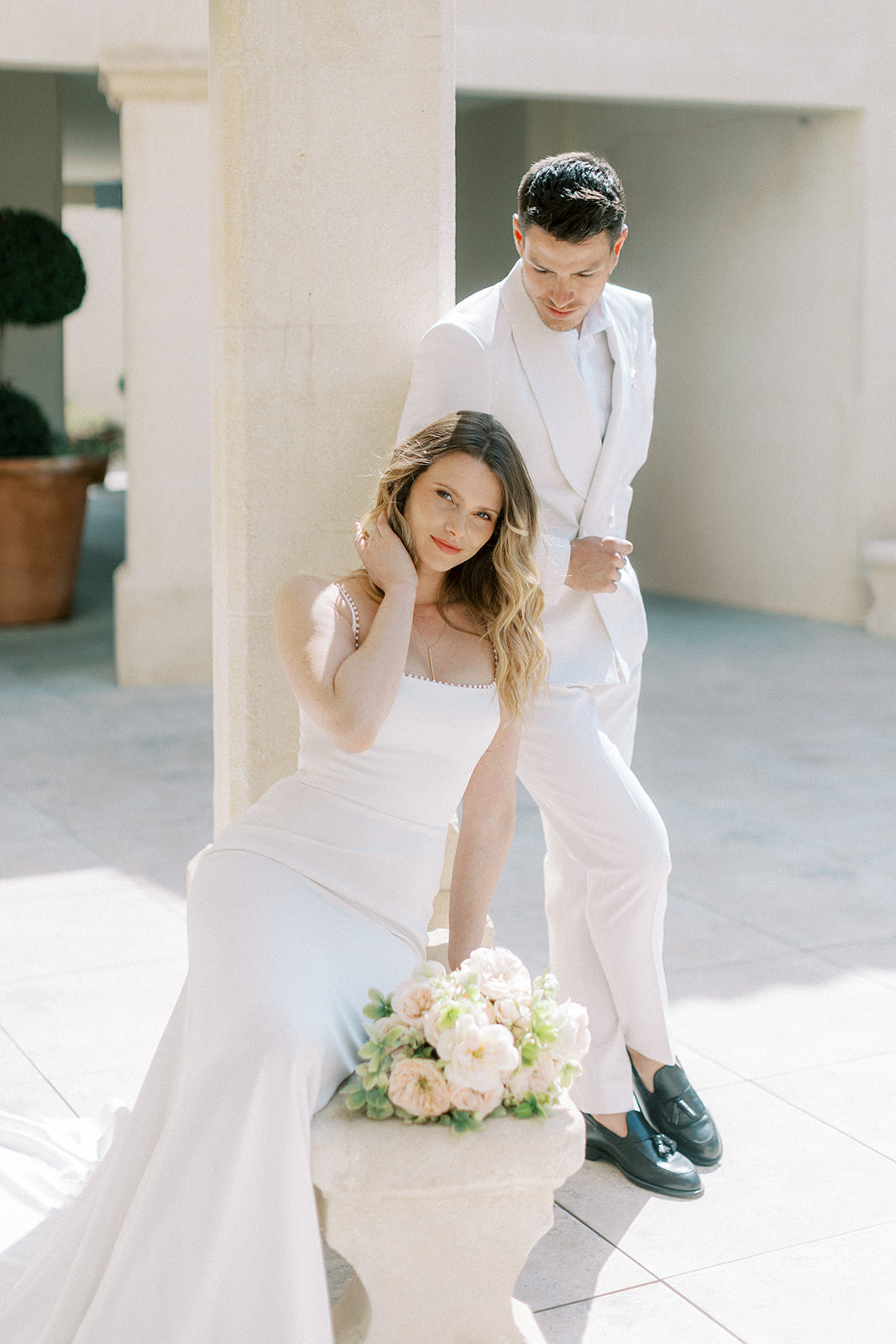 Bride in ivory slip gown and groom in white suit at colonnaded terrace with blush rose bouquet