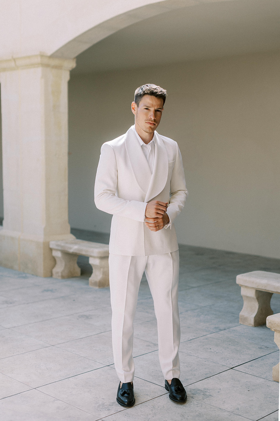 Groom in all-white tuxedo with shawl lapel adjusting cuffs on stone terrace with classical columns behind