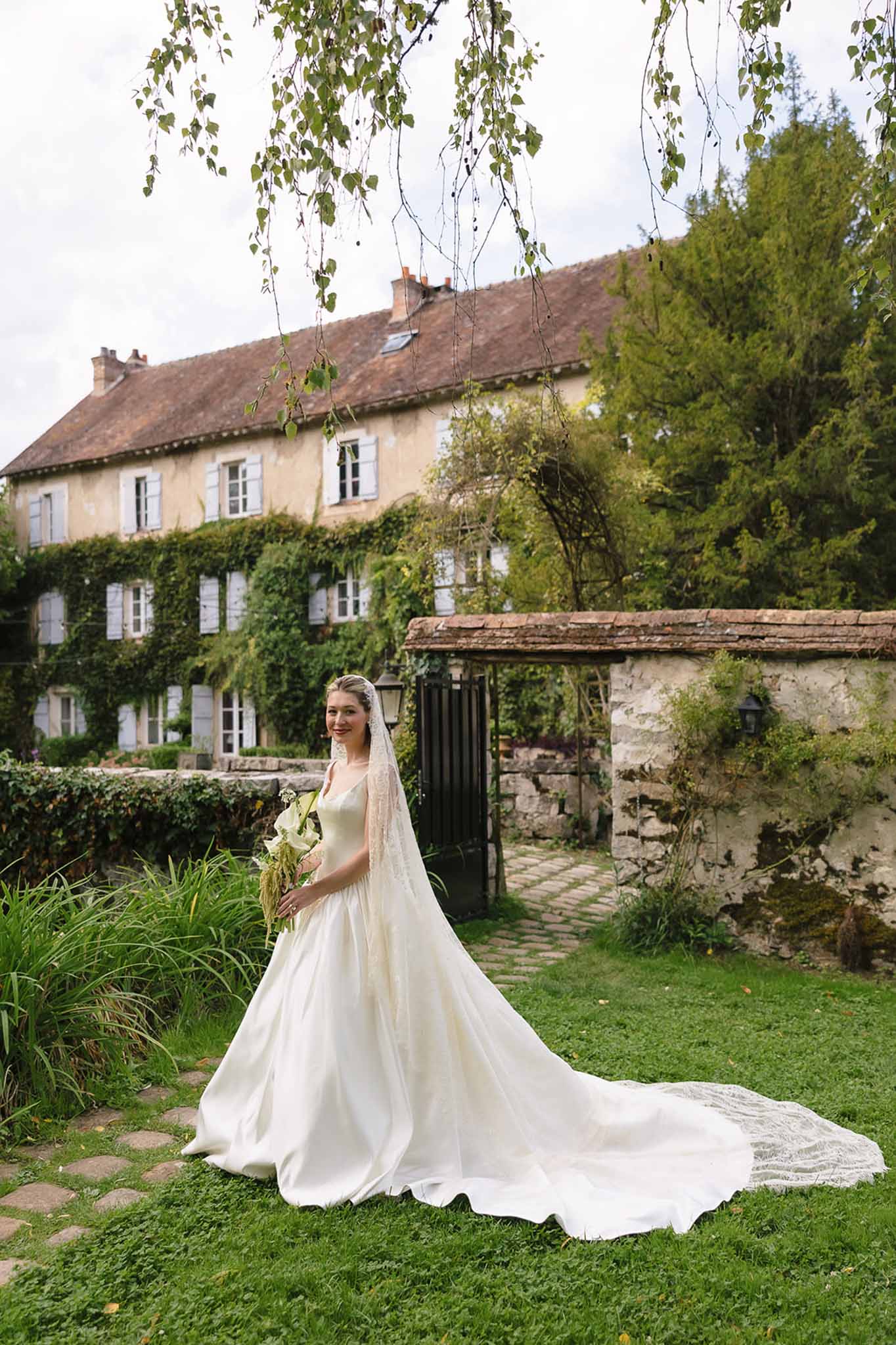 Bride in ivory satin ballgown with cathedral veil and calla lily bouquet standing on lawn before an ivy-covered French manor