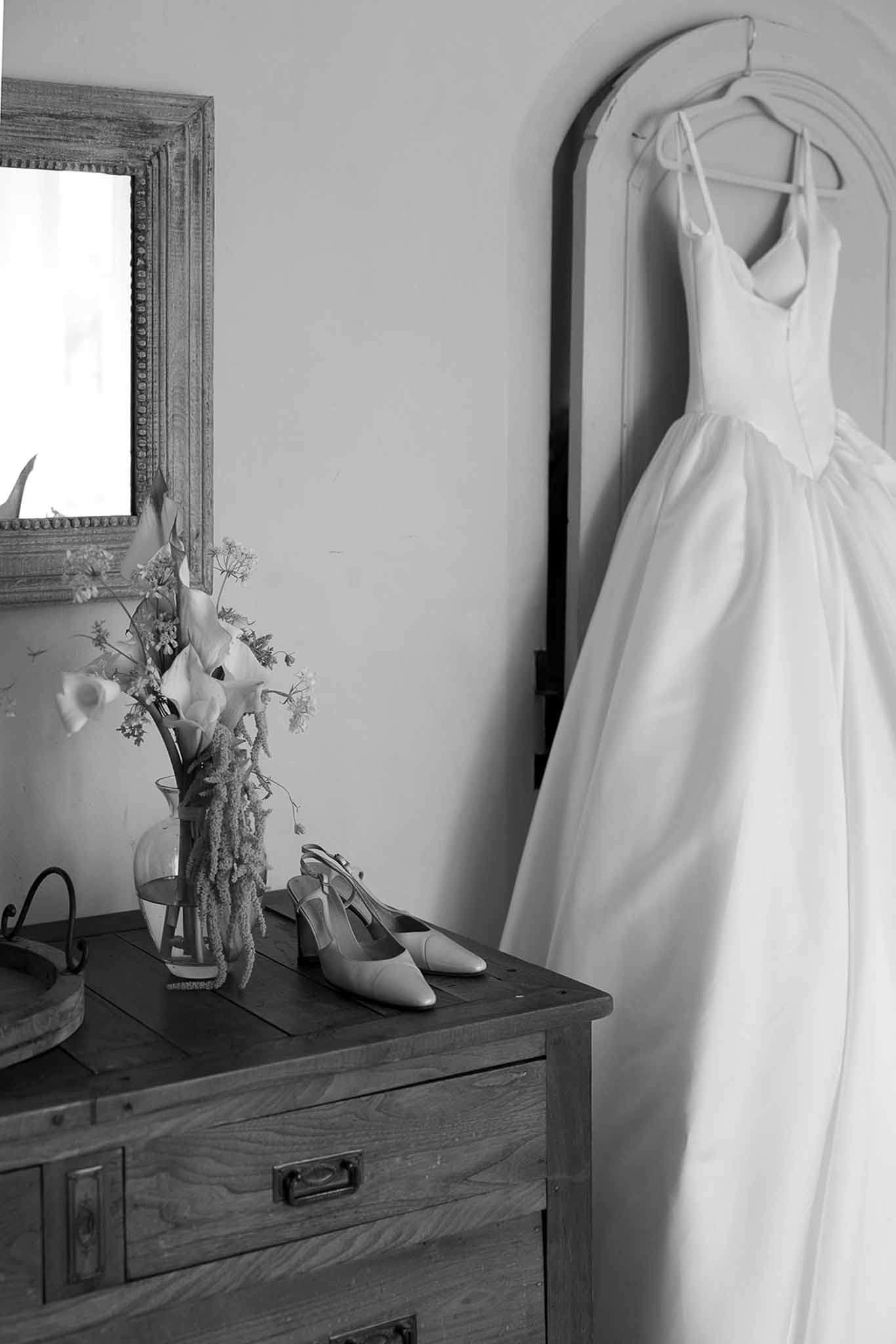Ballgown hanging in arched doorframe with calla lily arrangement and slingback heels on chest in B&W