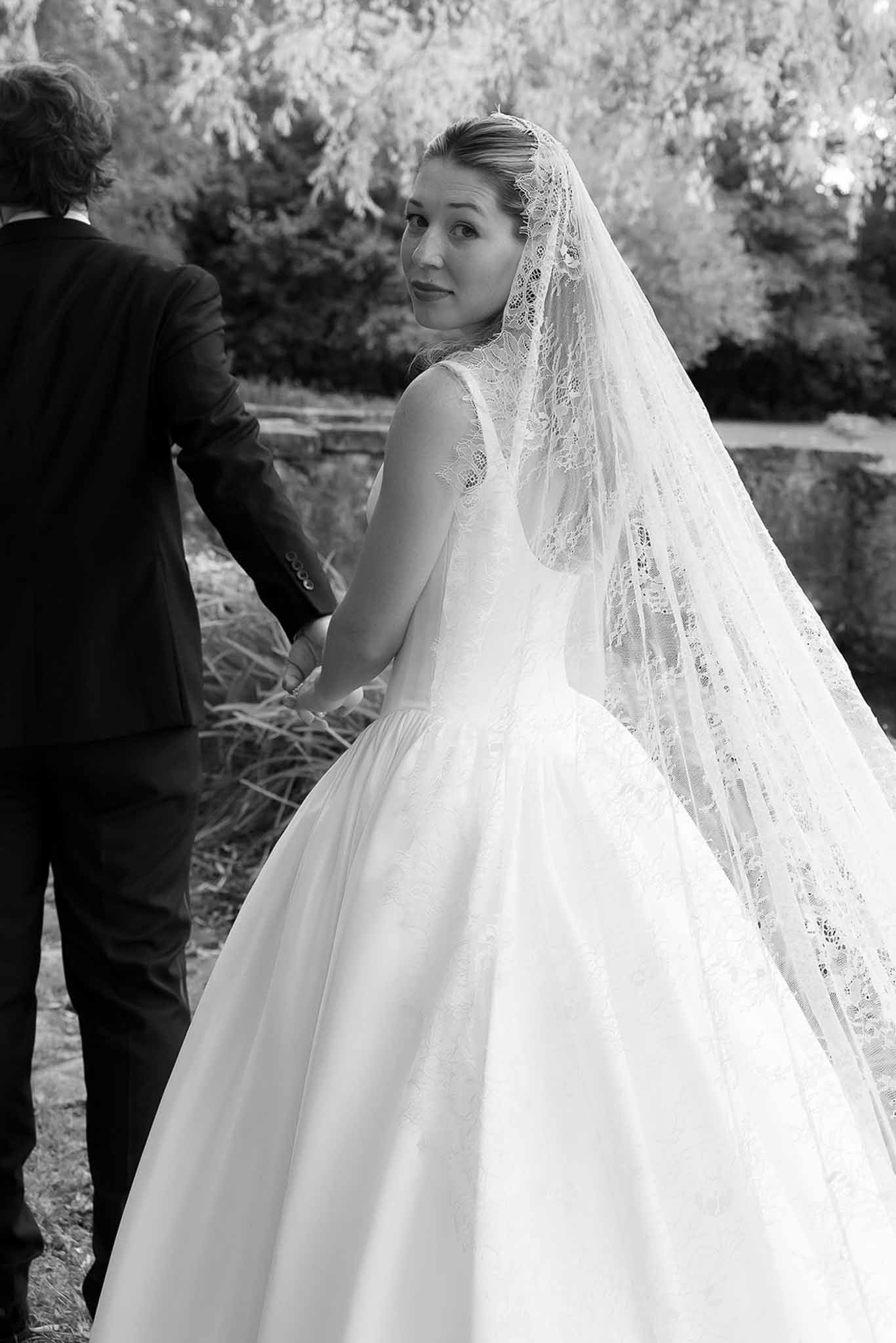 Bride in ballgown with lace-edged cathedral veil glancing back holding groom's hand in B&W garden portrait