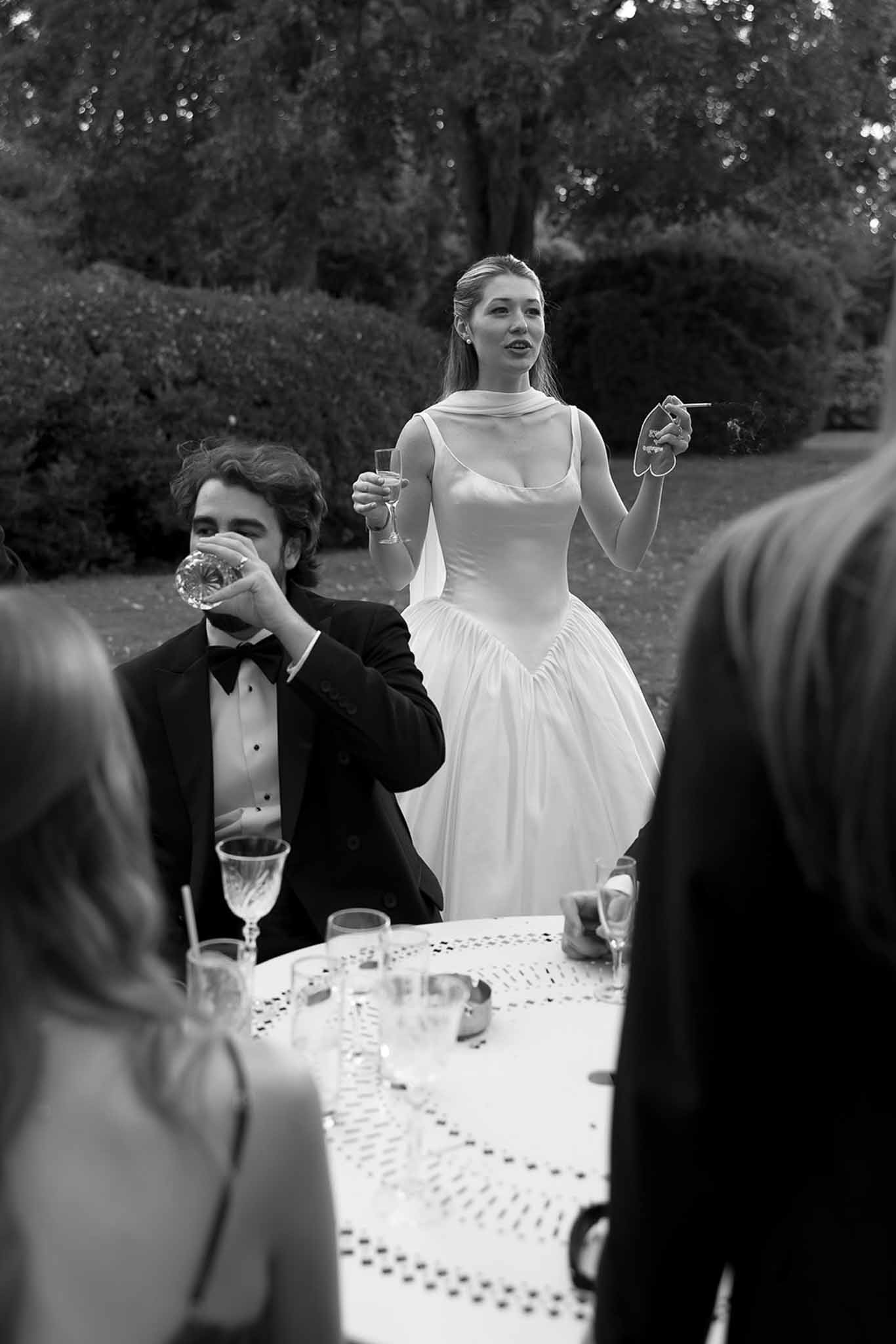Black and white image of bride giving speech at outdoor reception while guest drinks from crystal glass