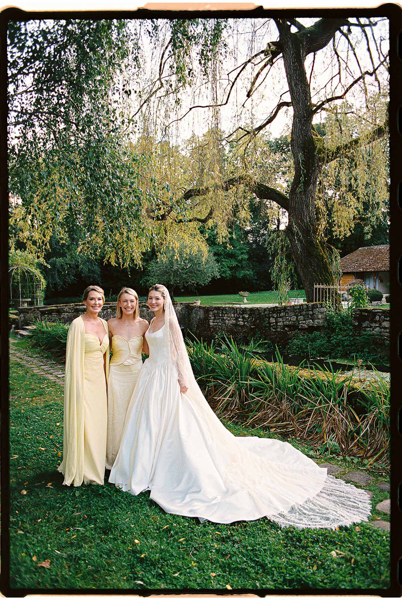 Bride in white satin ball gown with two bridesmaids in butter-yellow dresses posing in chateau garden