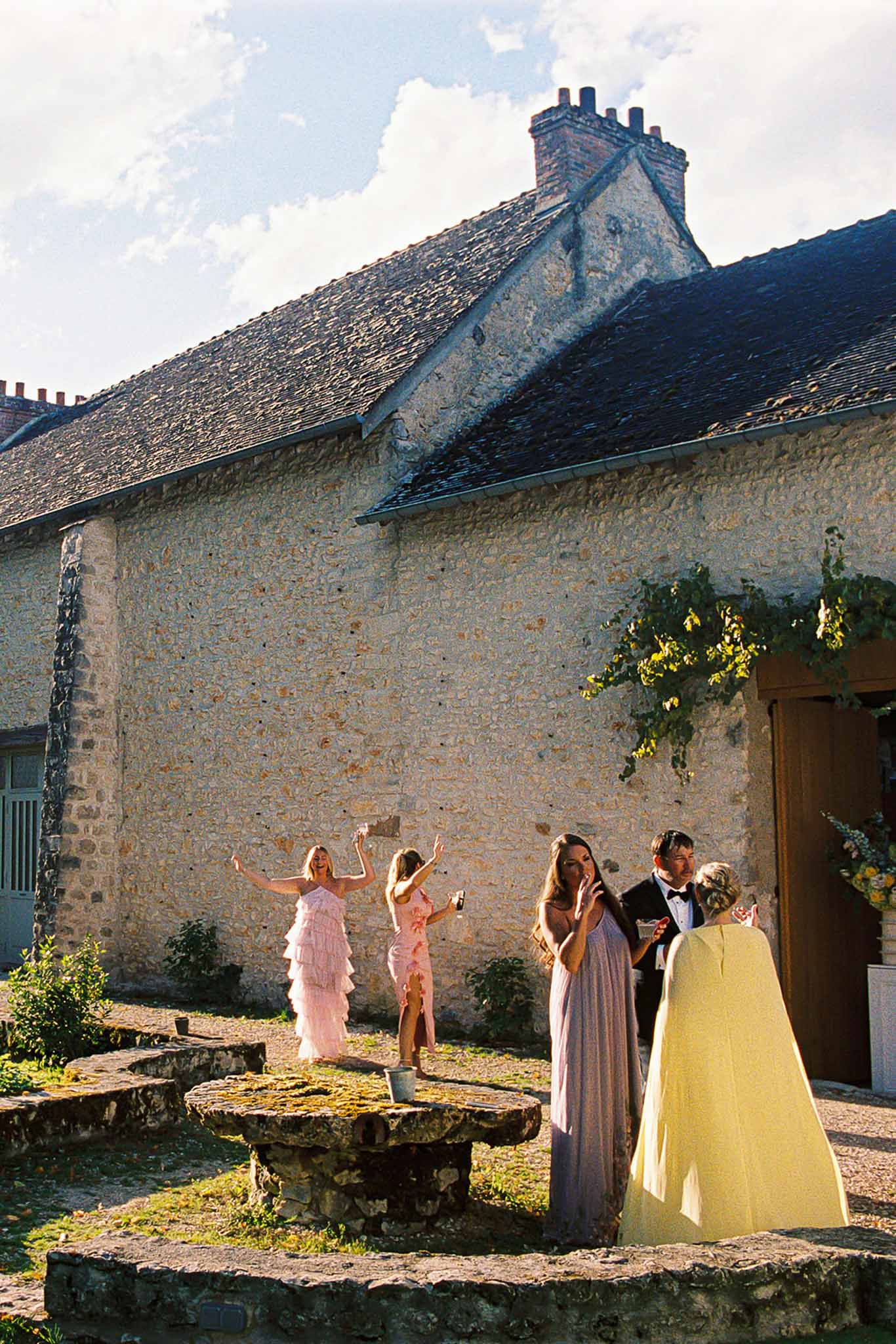 Guests dancing and socializing in courtyard of rustic French stone building during cocktail hour in afternoon sun