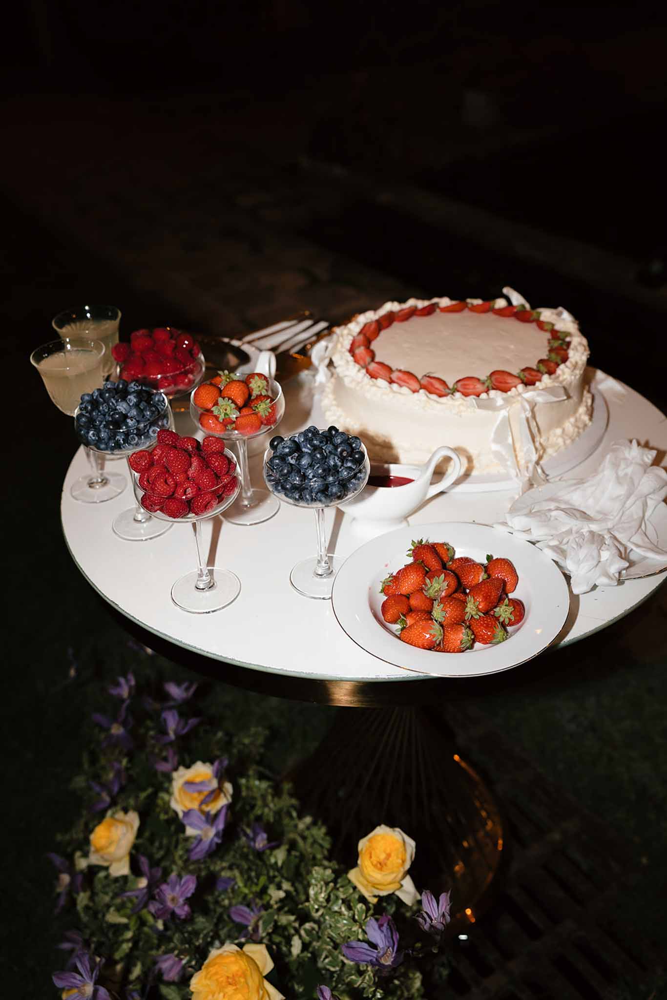 Wedding cake table with single-tier white cake topped with strawberries, coupe glasses of fresh berries, and yellow roses