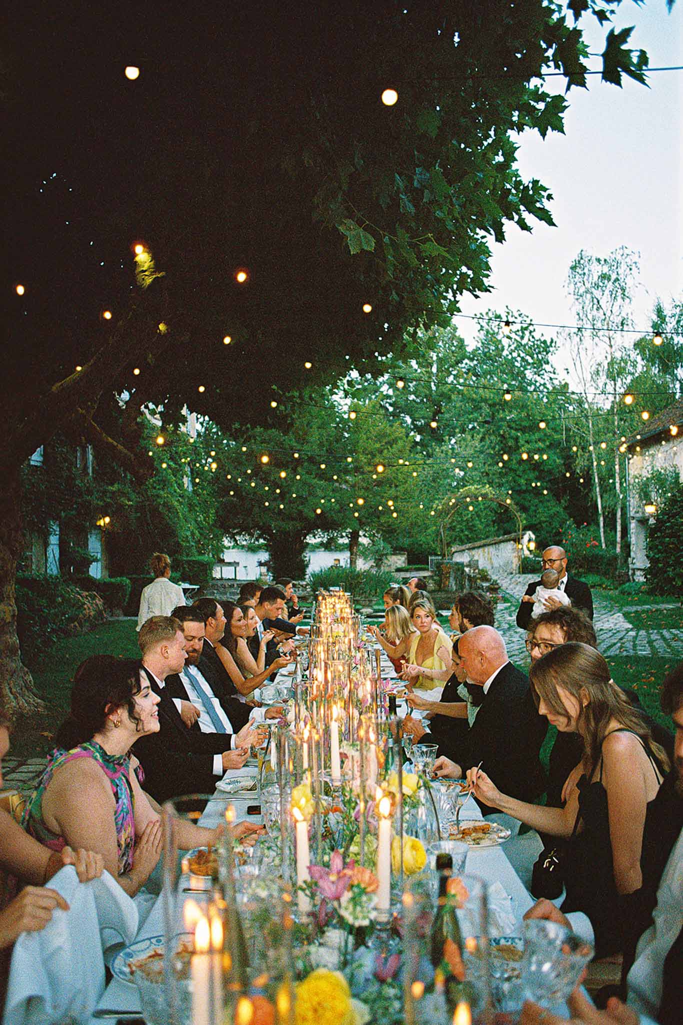 Outdoor wedding reception dinner at dusk with guests seated at long banquet table with floral runner and taper candles