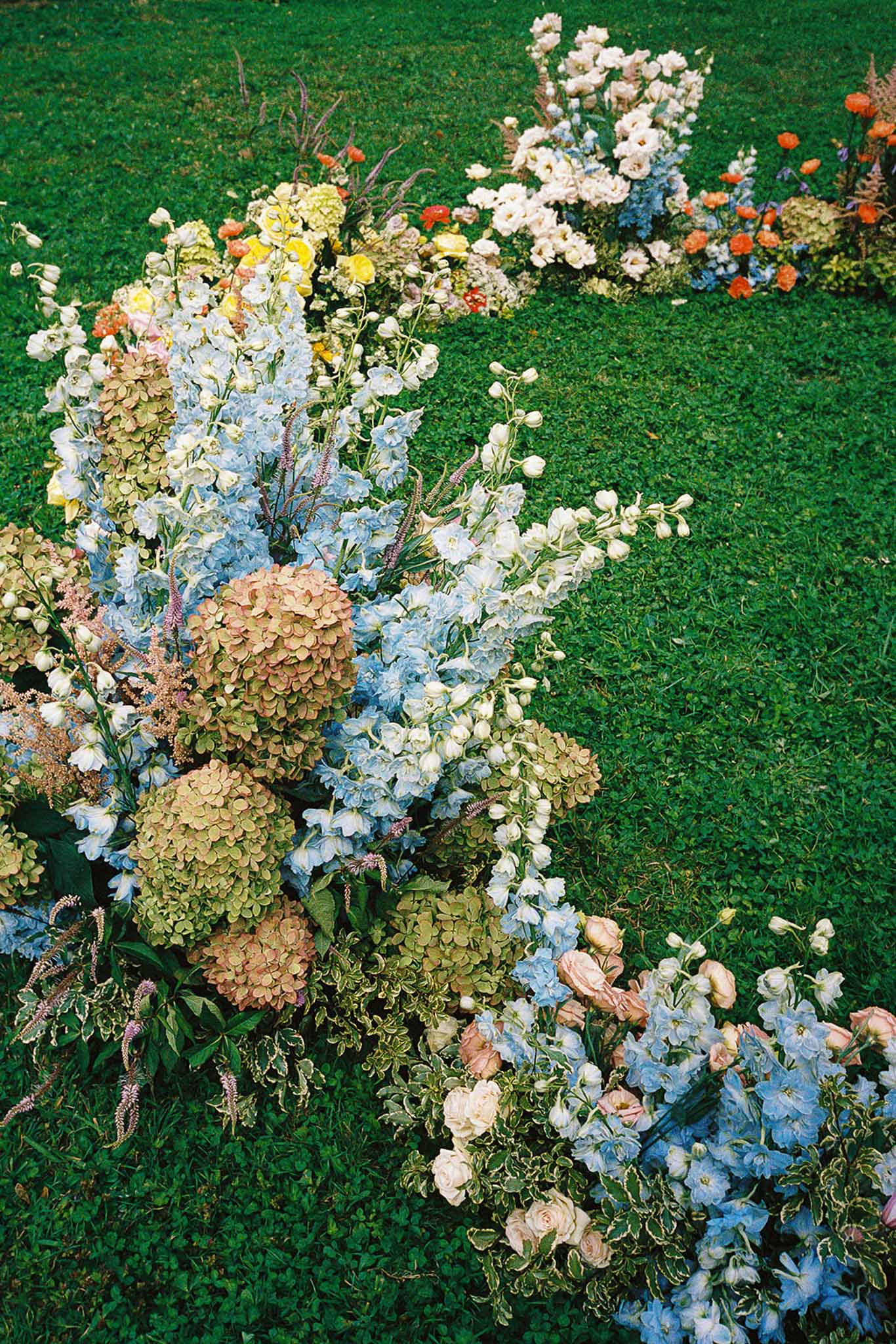 Ground-level garden floral installation with blue delphiniums, blush roses, and purple veronica on lawn