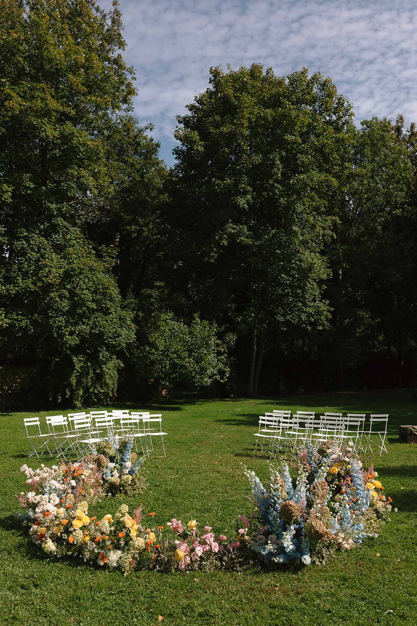 Garden lawn ceremony setup with white metal chairs and colorful floral ground installations framing the aisle
