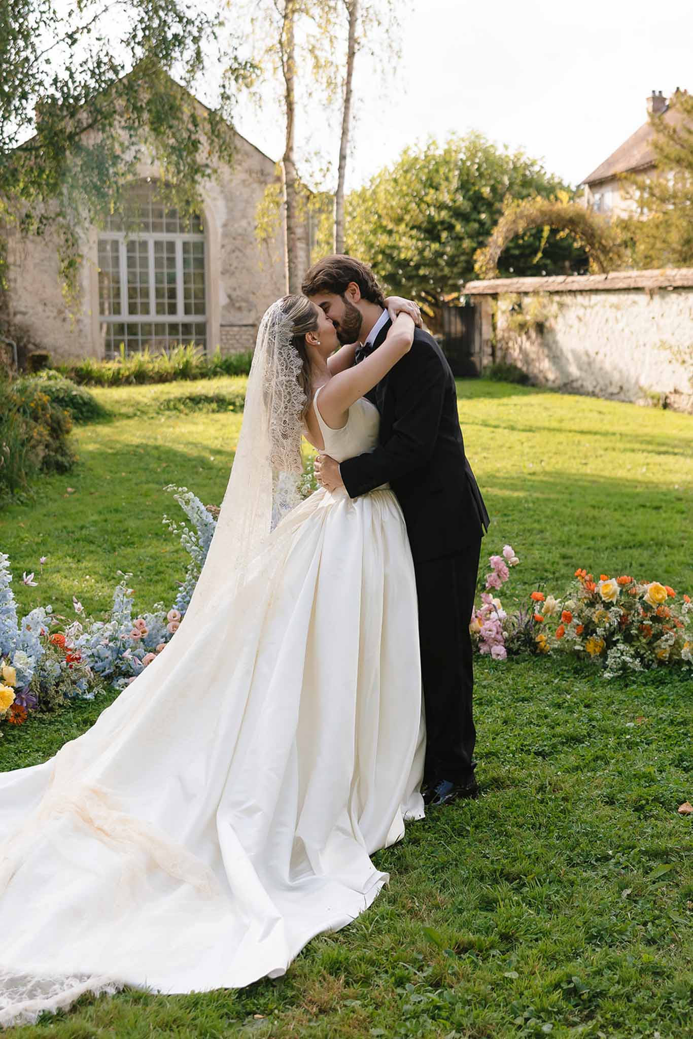 Bride in satin ballgown and groom in black tuxedo kissing in garden surrounded by colorful floral clusters