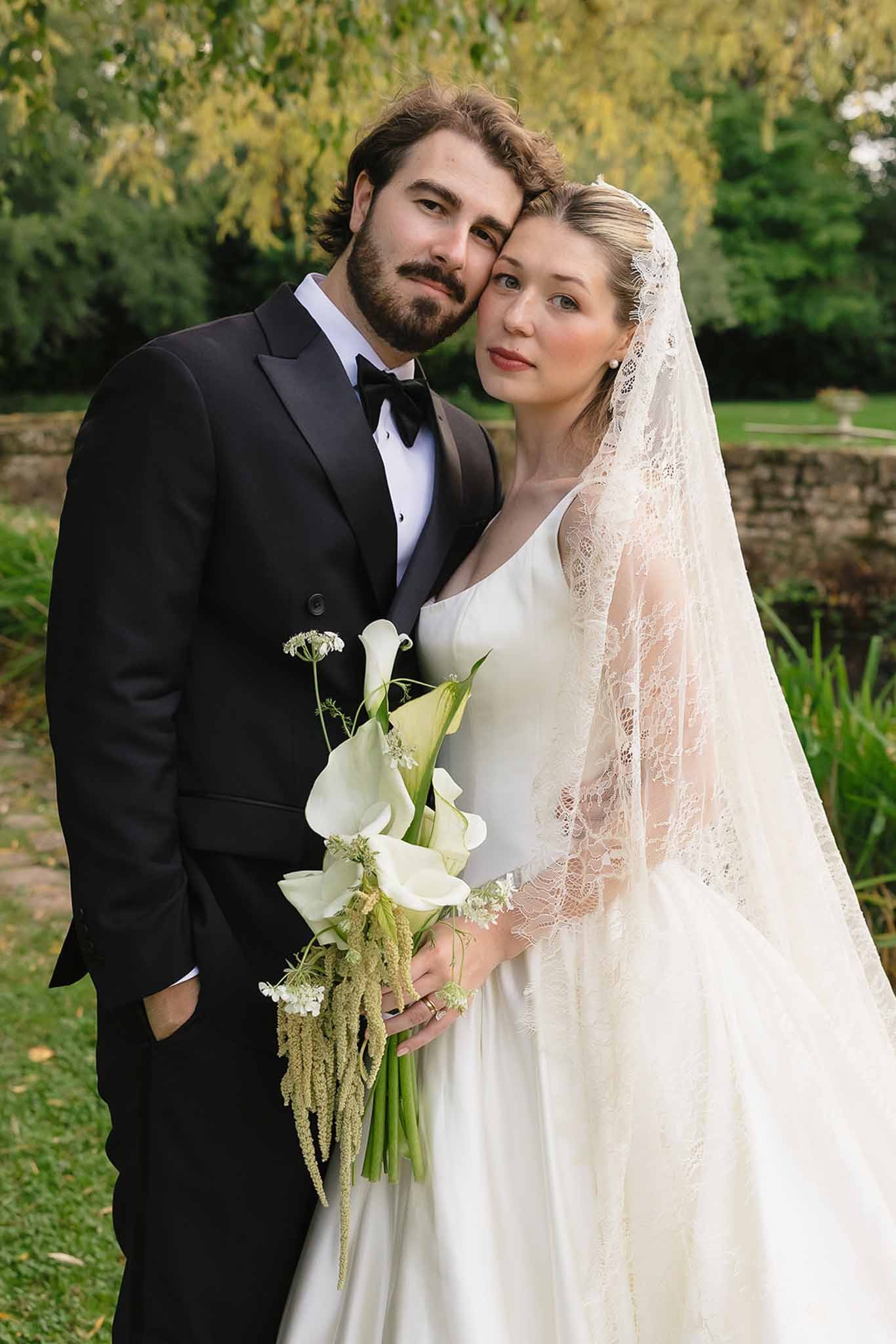 Close-up of bride in lace-edged veil holding white calla lily bouquet leaning against groom in black tuxedo