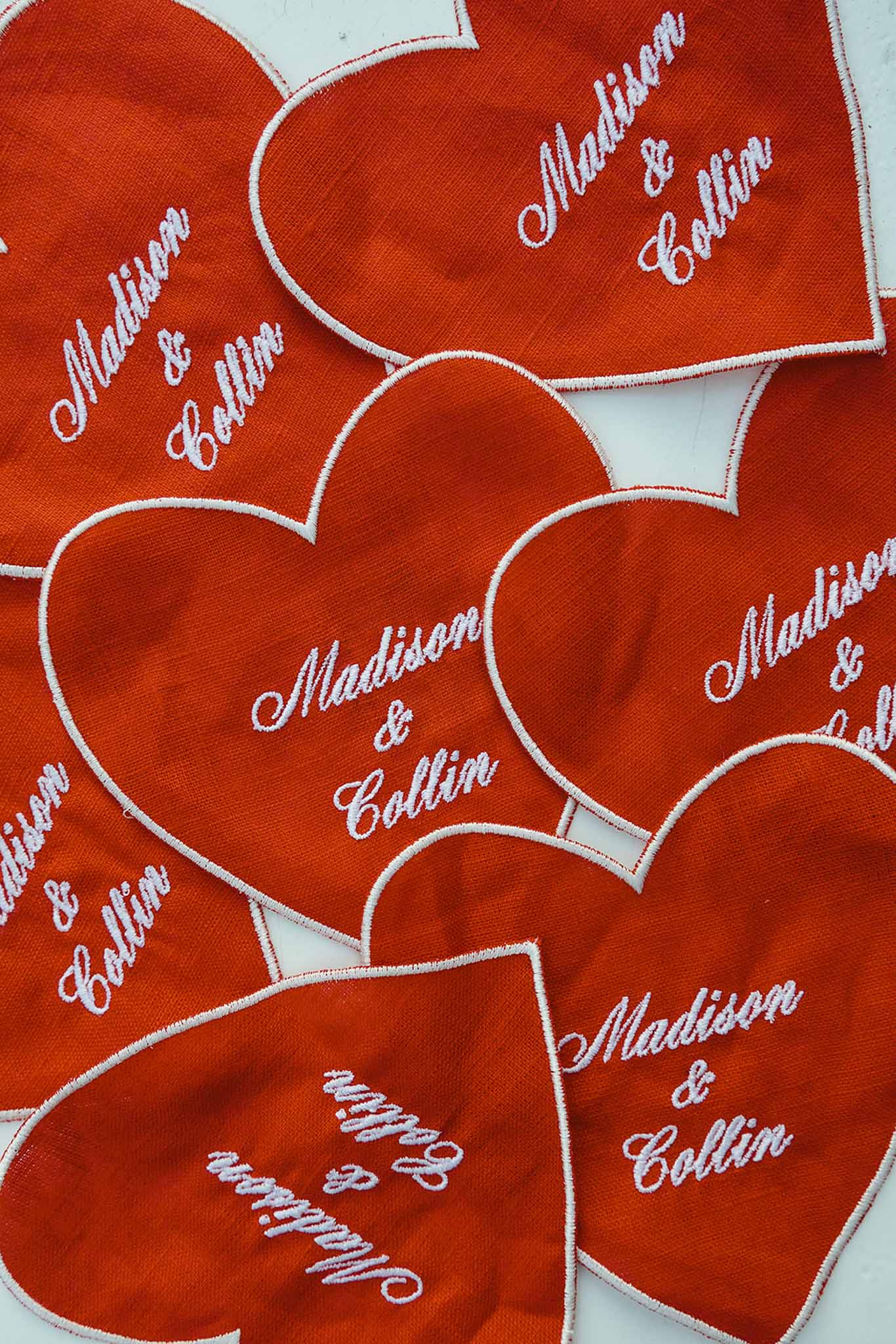 Heart-shaped embroidered patches in burnt orange with white border reading Madison and Collin scattered on white surface