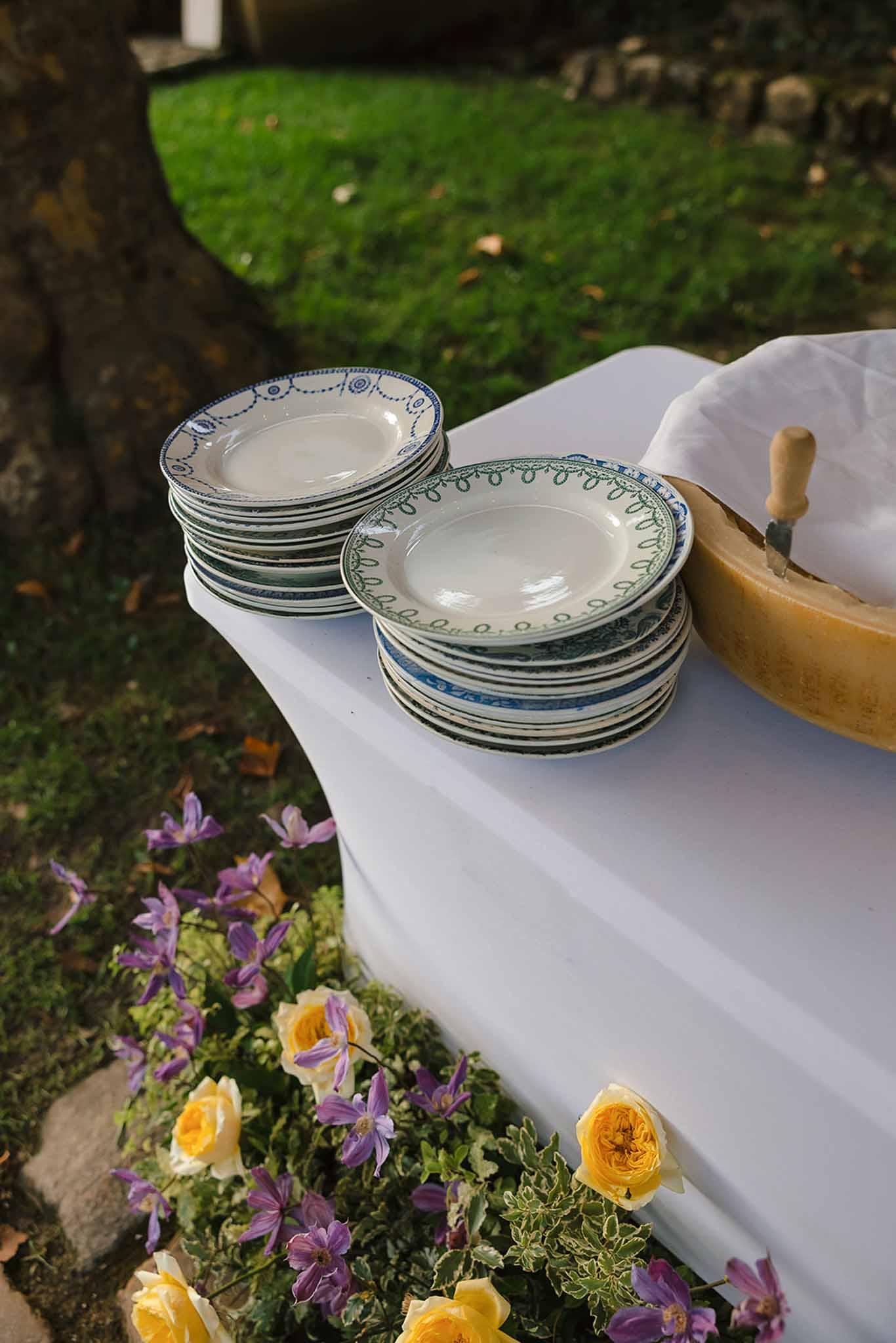 Vintage mismatched plates with blue and green borders beside cheese wheel and yellow roses on white linen