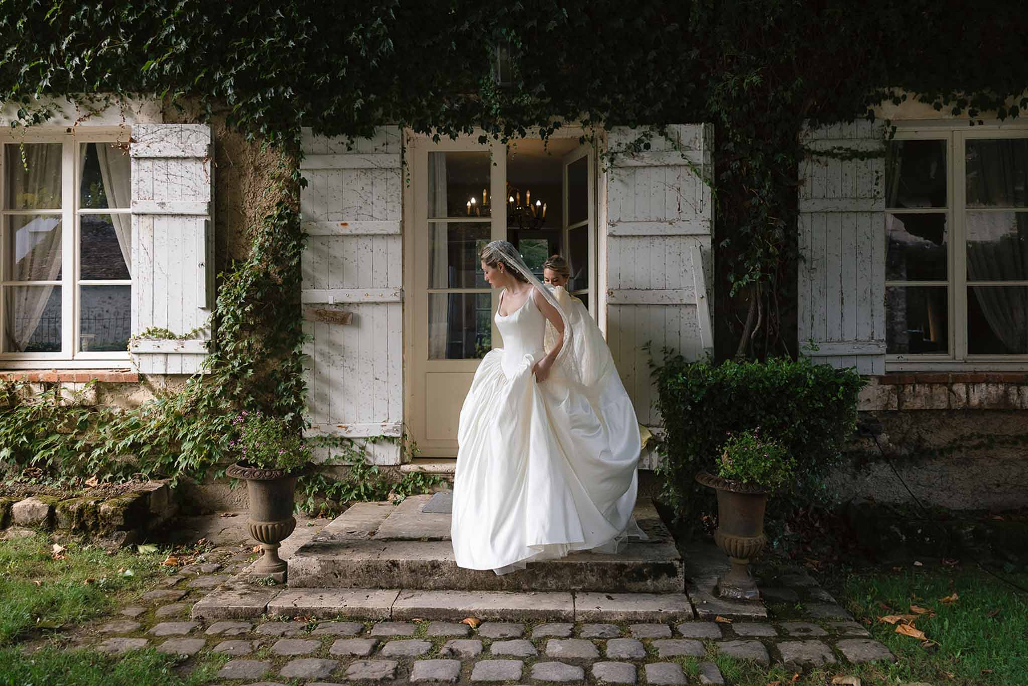 Bride in ivory satin ballgown stepping down stone stairs at French manor with ivy-covered walls