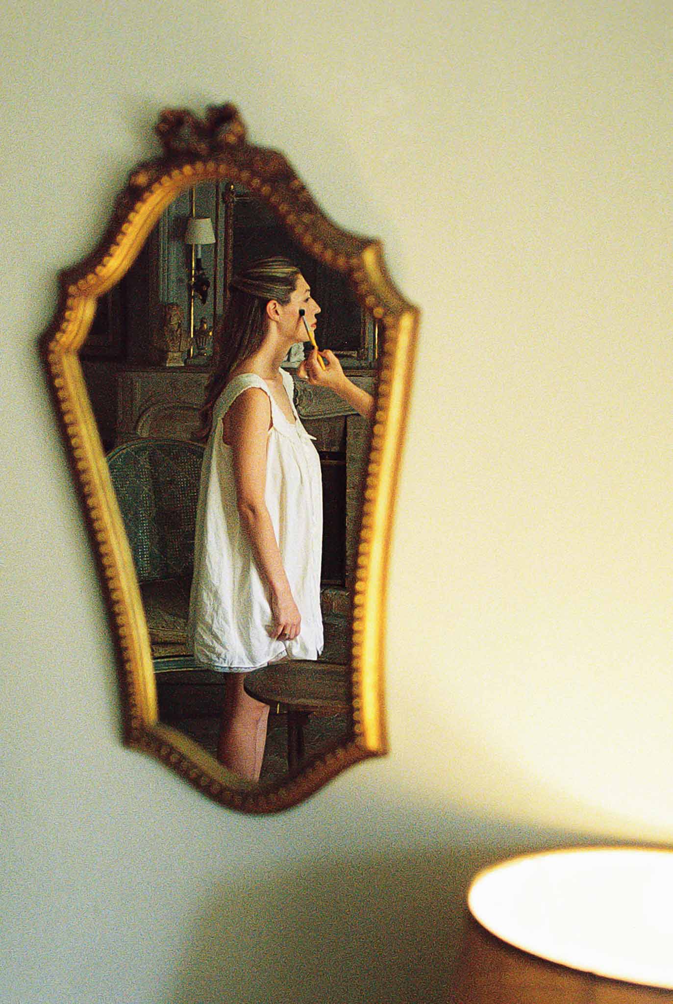 Bride applying makeup reflected in ornate gold-framed crown mirror with antique furnishings in warm film-grain light