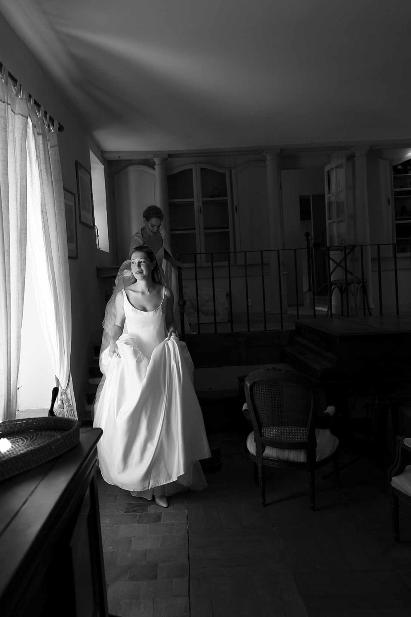 Bride in spaghetti-strap gown as attendant arranges veil on mezzanine in Provencal interior in B&W