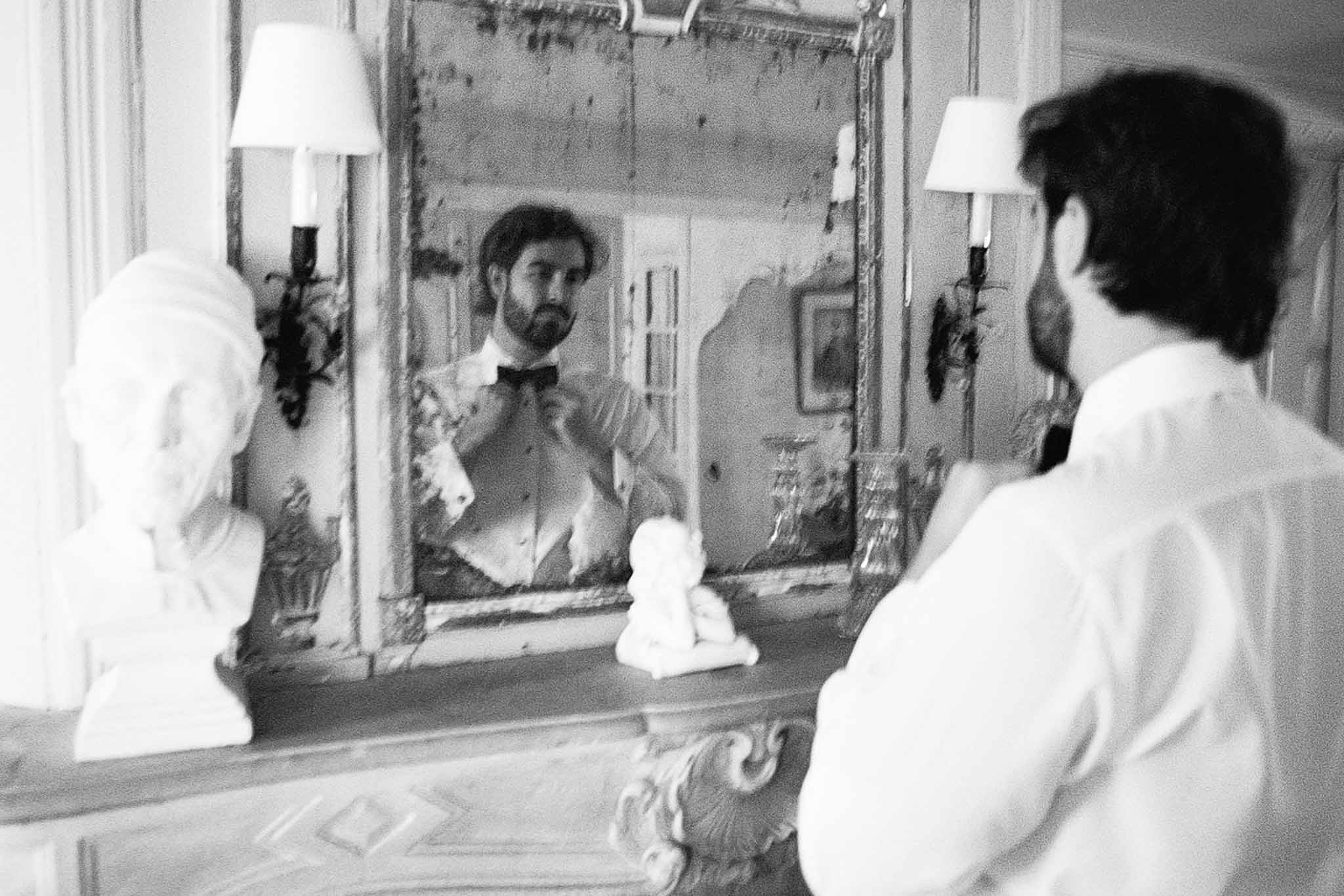 Black and white shot of groom adjusting bow tie in ornate gilt-framed mirror in classic French chateau interior