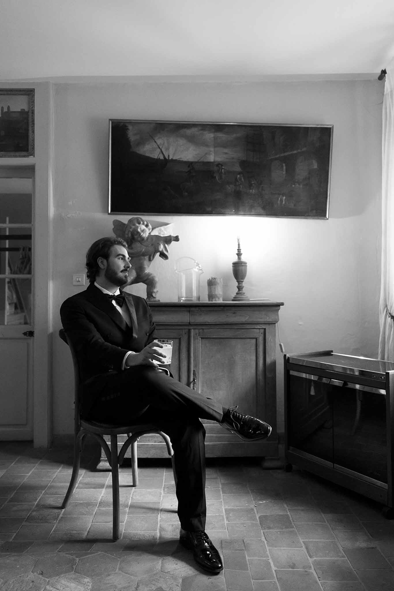 Groom in tuxedo sits with drink beside vintage sideboard with cherub and landscape painting in B&W