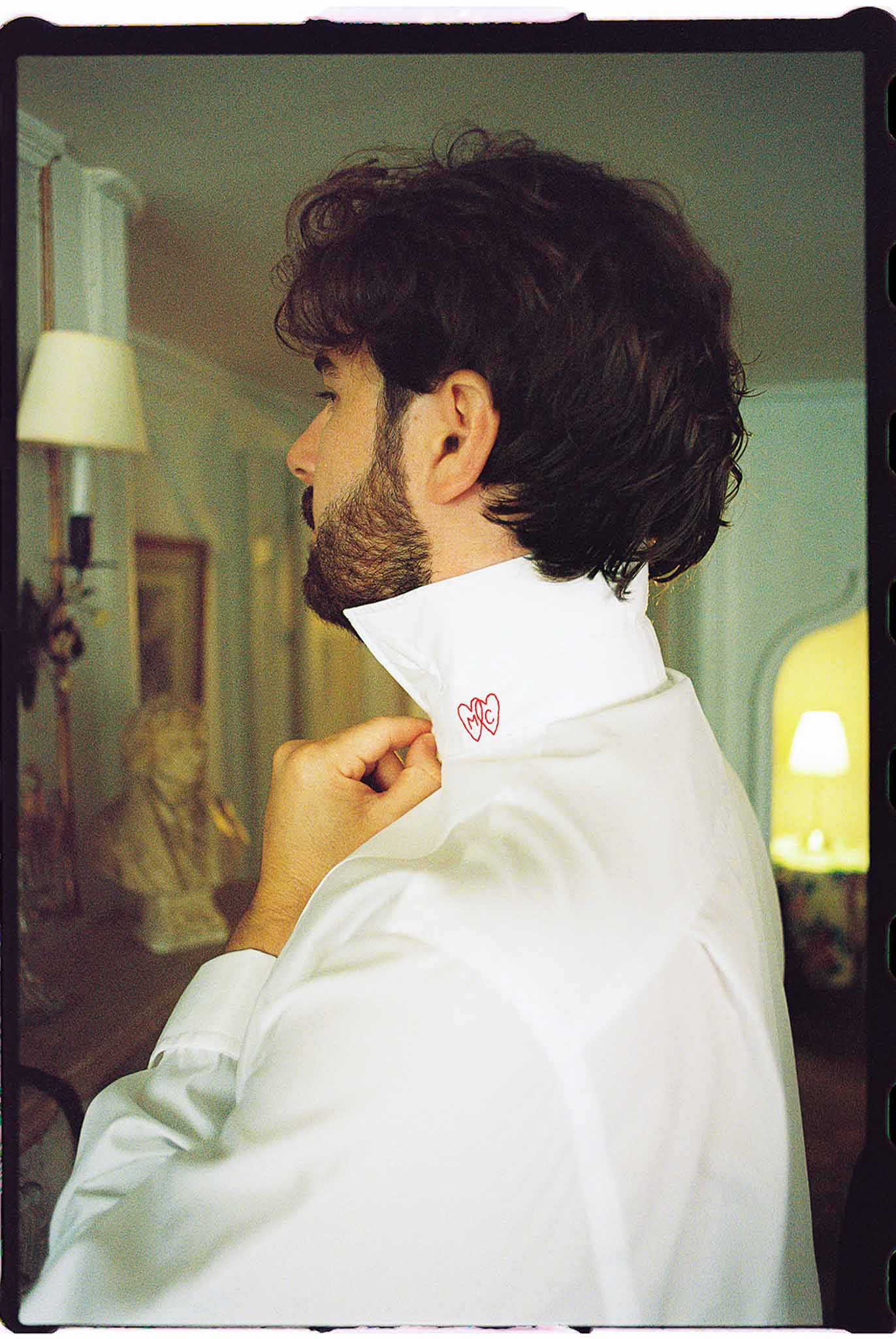 Groom buttoning white dress shirt with custom red embroidered monogram initials in a chateau room
