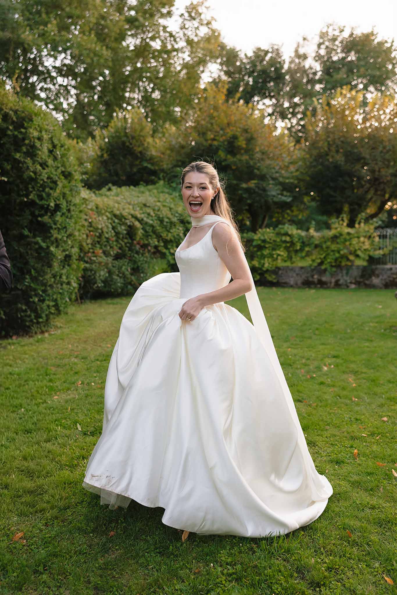 Bride laughing and gathering her ivory satin ball gown skirt on a manicured garden lawn in late afternoon light