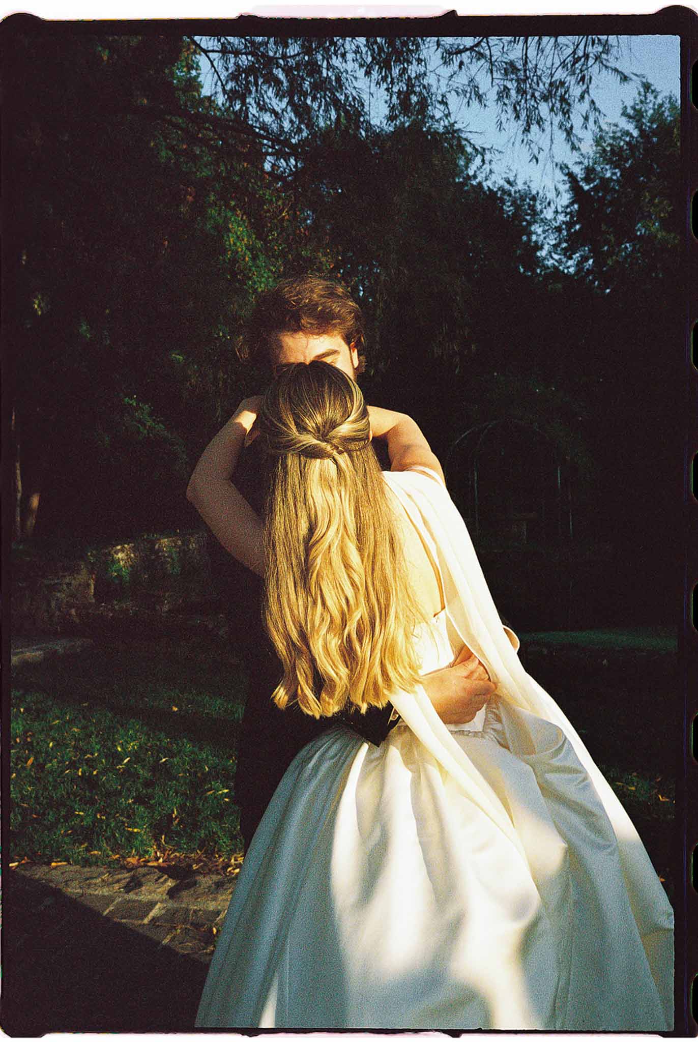 Bride and groom embrace in garden at golden hour, bride in ivory ball gown with dark sash, warm backlight