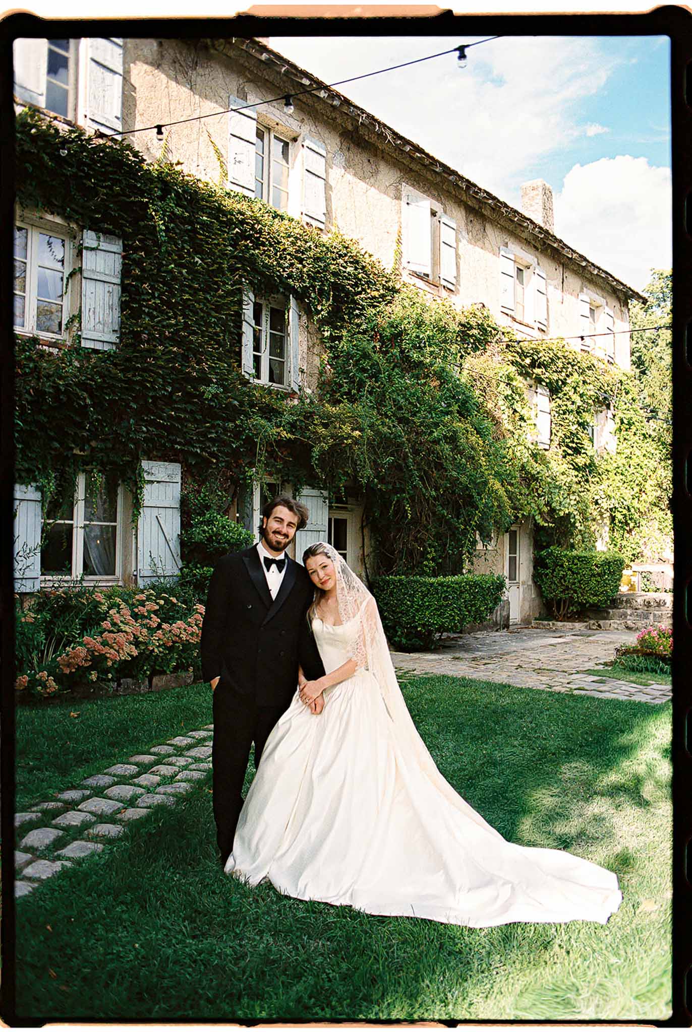 Film portrait of bride leaning on groom before ivy-covered mas with blue shutters and string lights