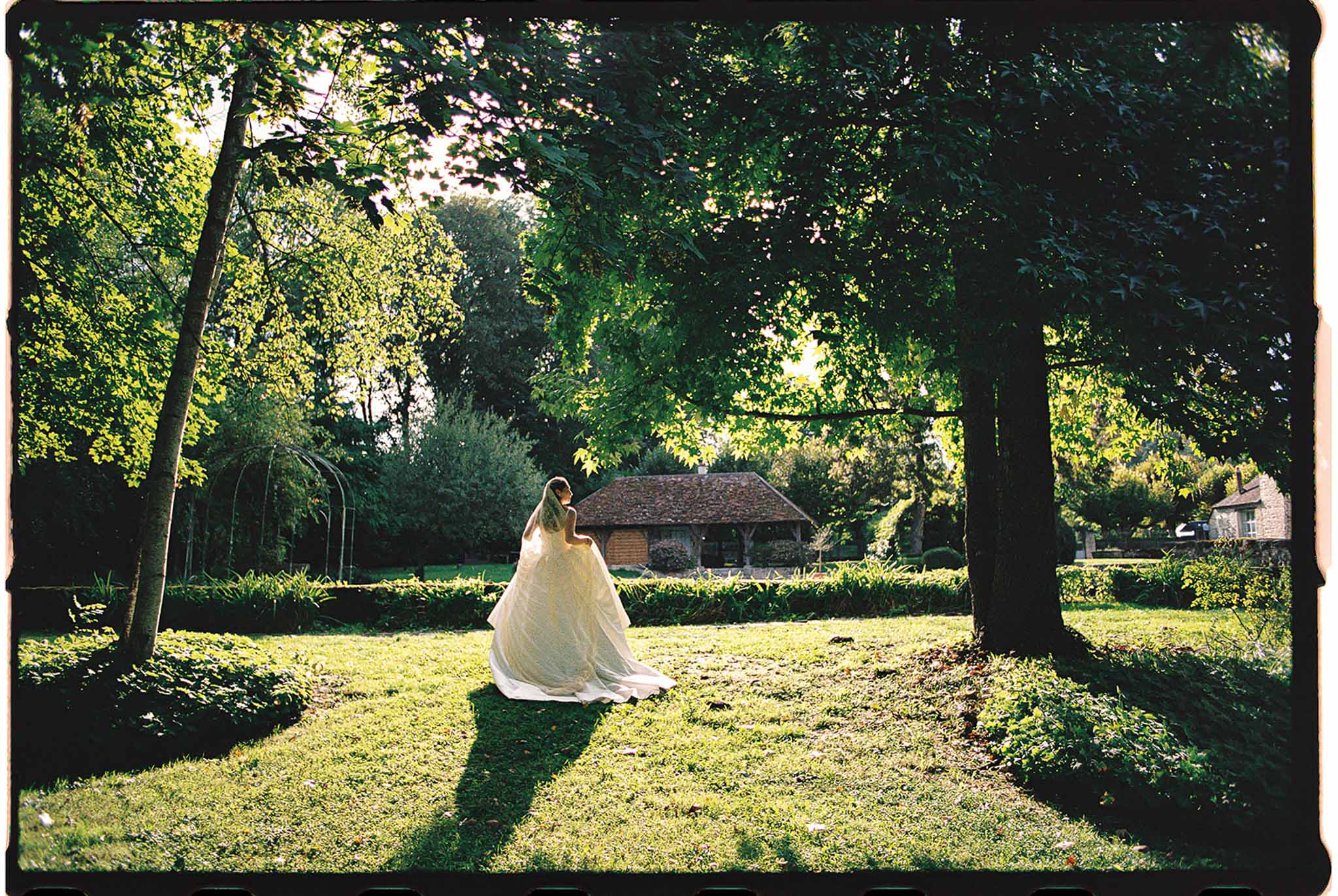 Bride in ivory ball gown and cathedral veil photographed from behind on a French estate lawn with mature trees