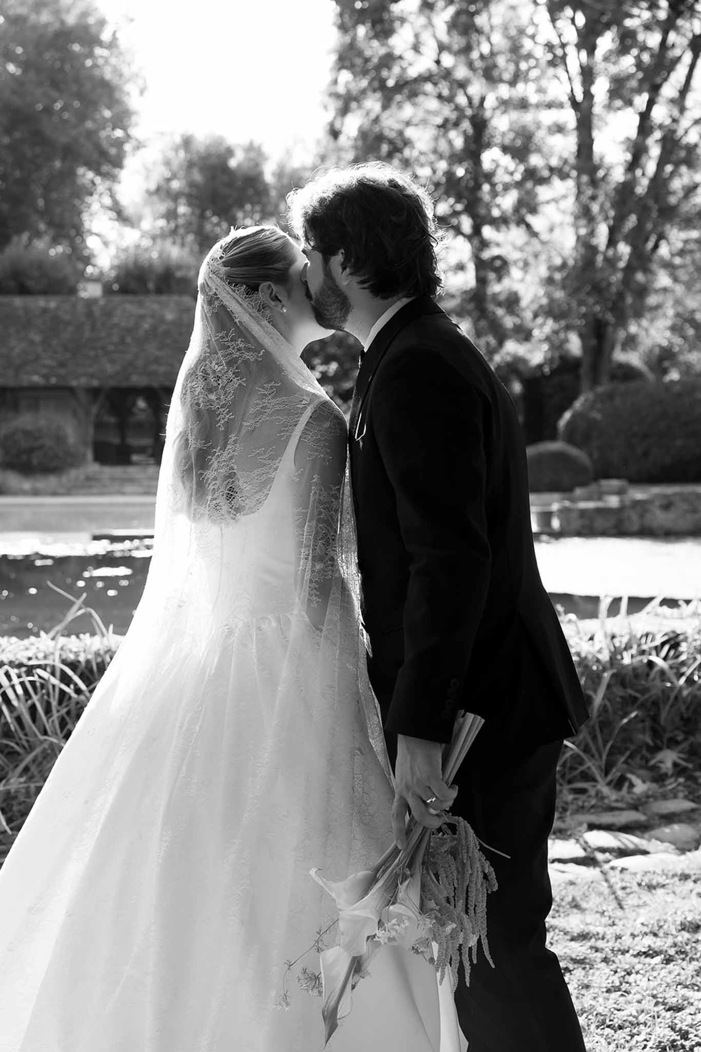 Black-and-white portrait of bride and groom kissing in formal garden with ornamental pond and topiary