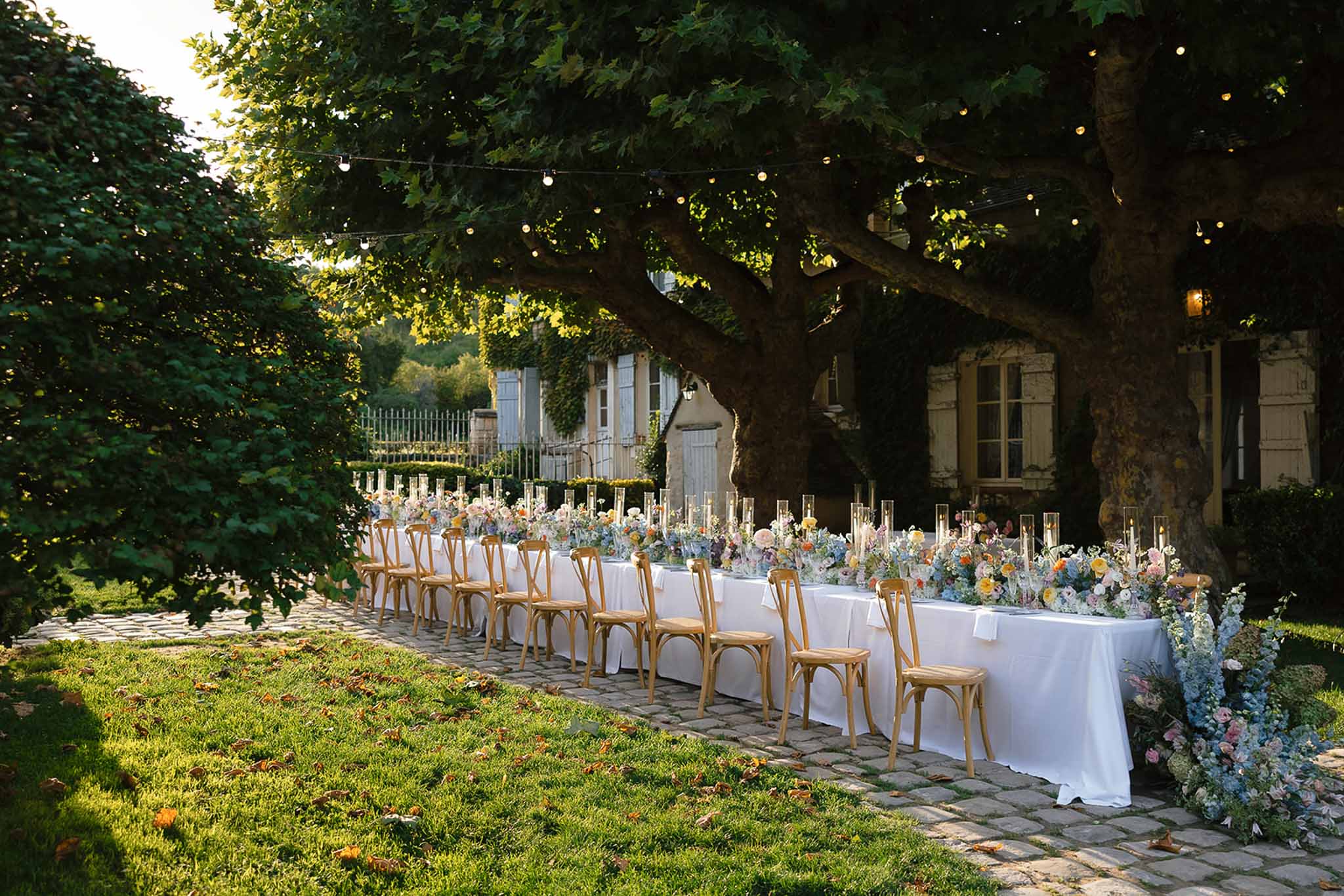 Long table with blue hydrangea and peach rose runner under fairy-lit tree on cobblestone path before stone manor