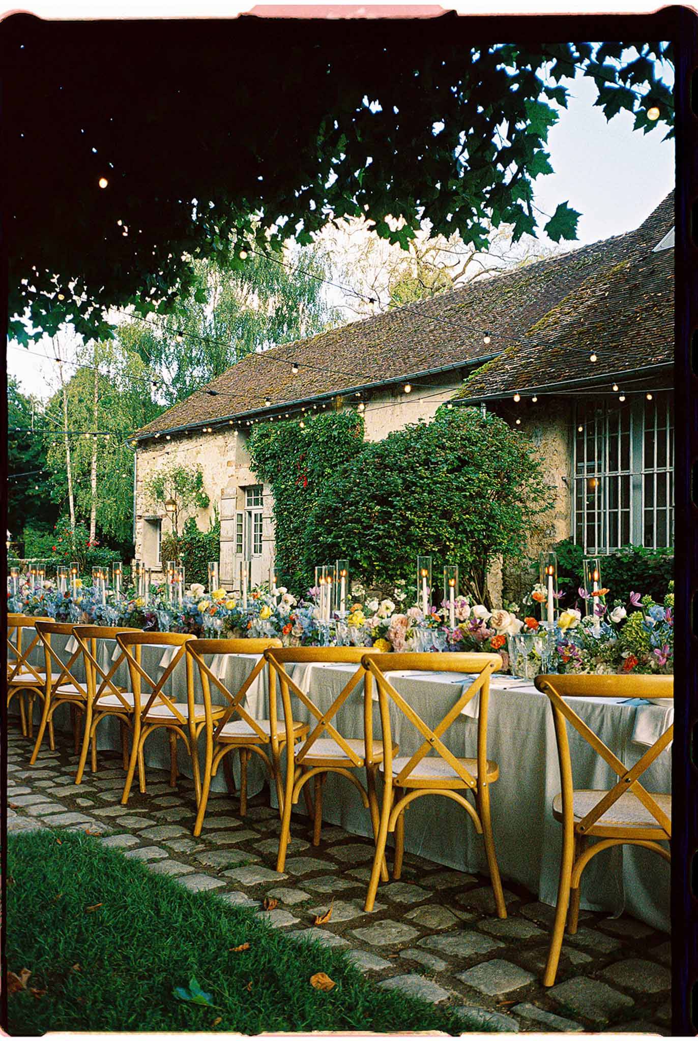 Farm tables with multicolour floral runners and hurricane candles under string lights at ivy-covered stone farmhouse