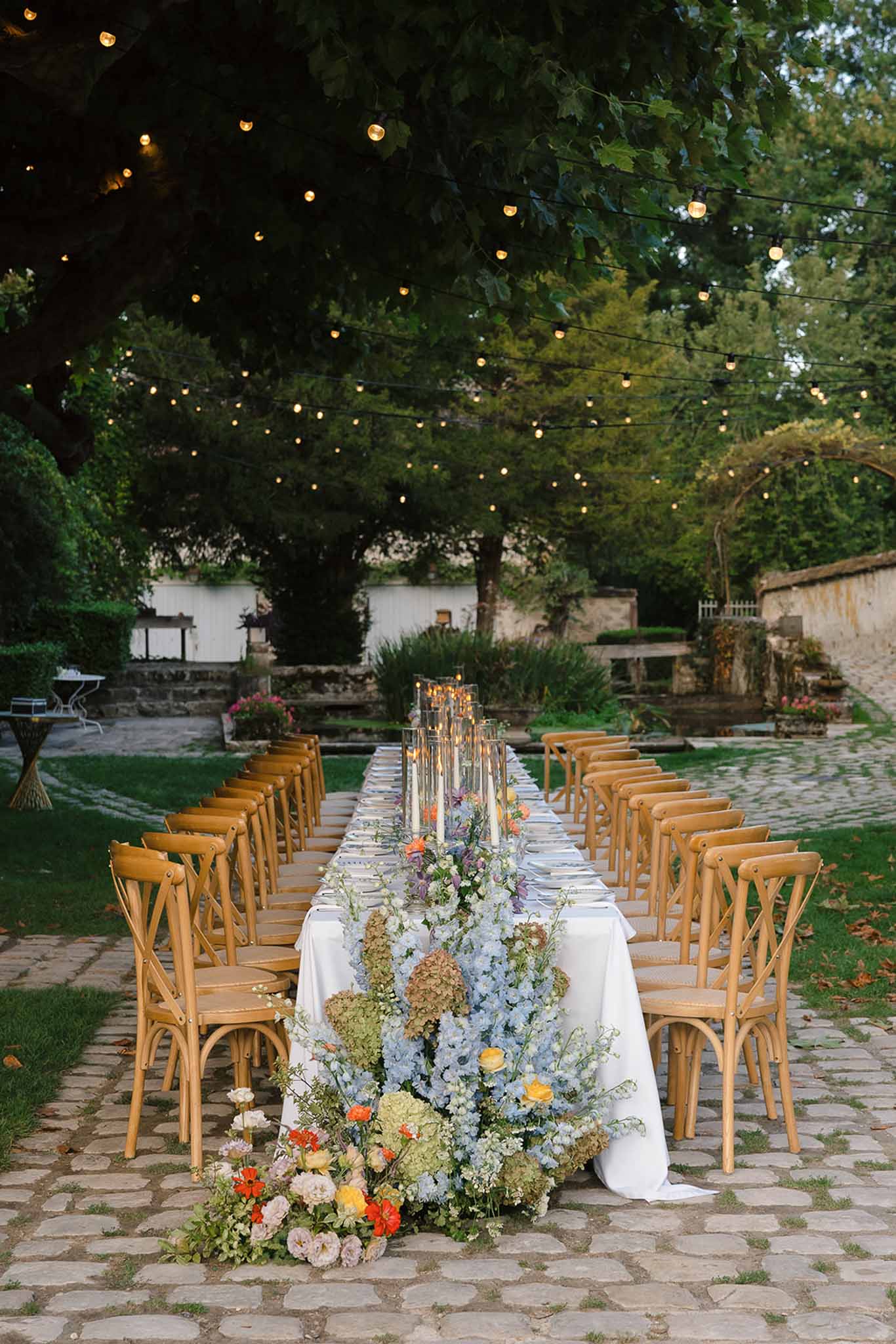Long outdoor reception table with blue delphiniums, blush roses, and orange ranunculus under string lights at dusk