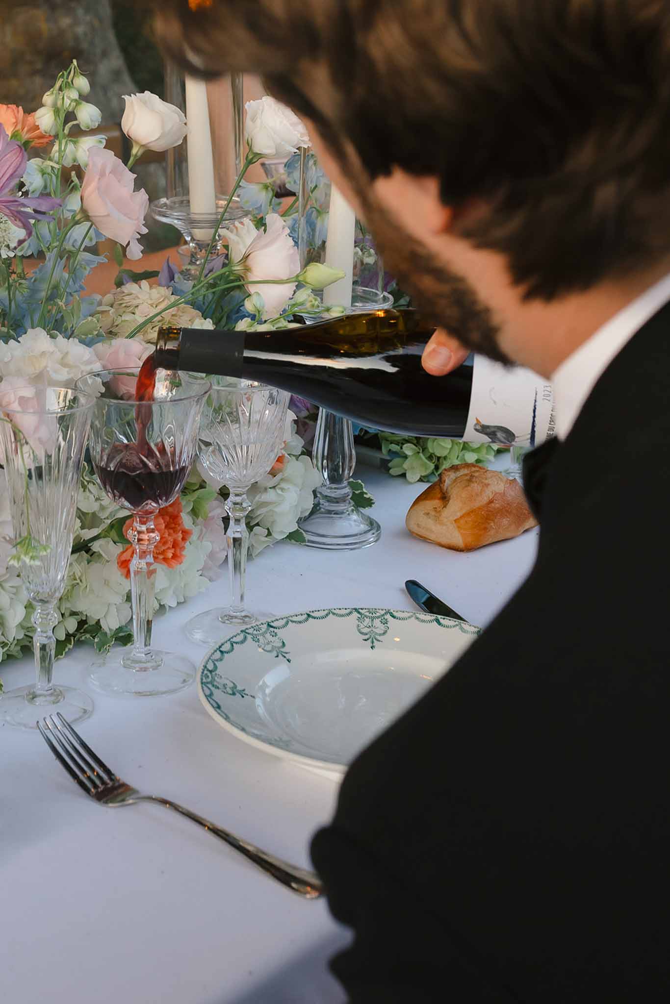 Guest pouring red wine beside blush rose and blue delphinium centerpiece with crystal stemware and china