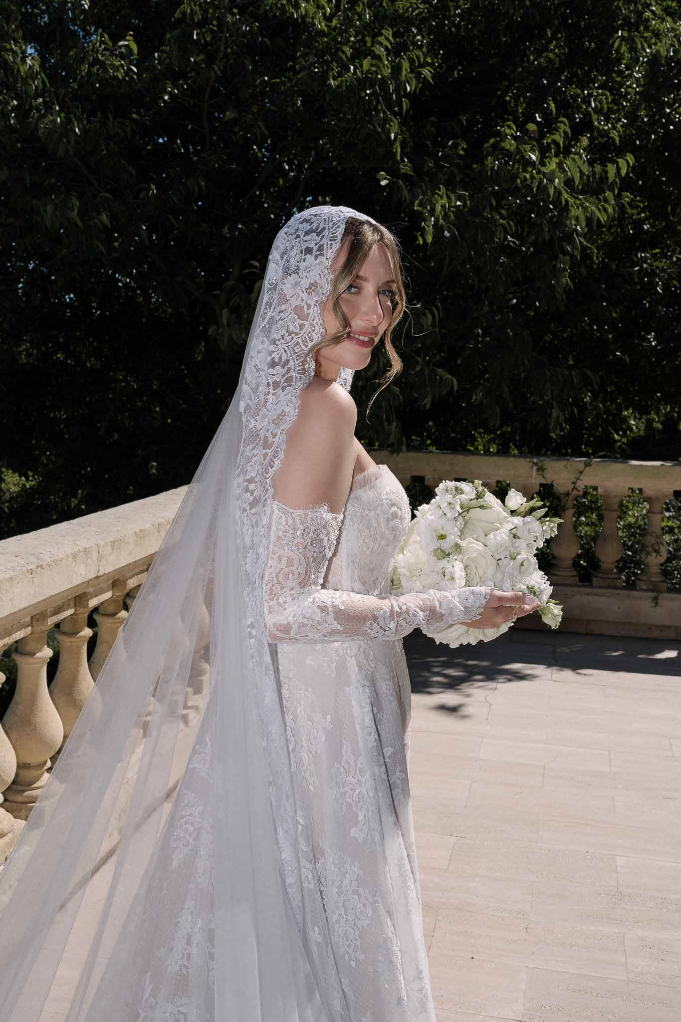 Bride in embroidered lace gown with mantilla veil holding white ranunculus bouquet on stone terrace