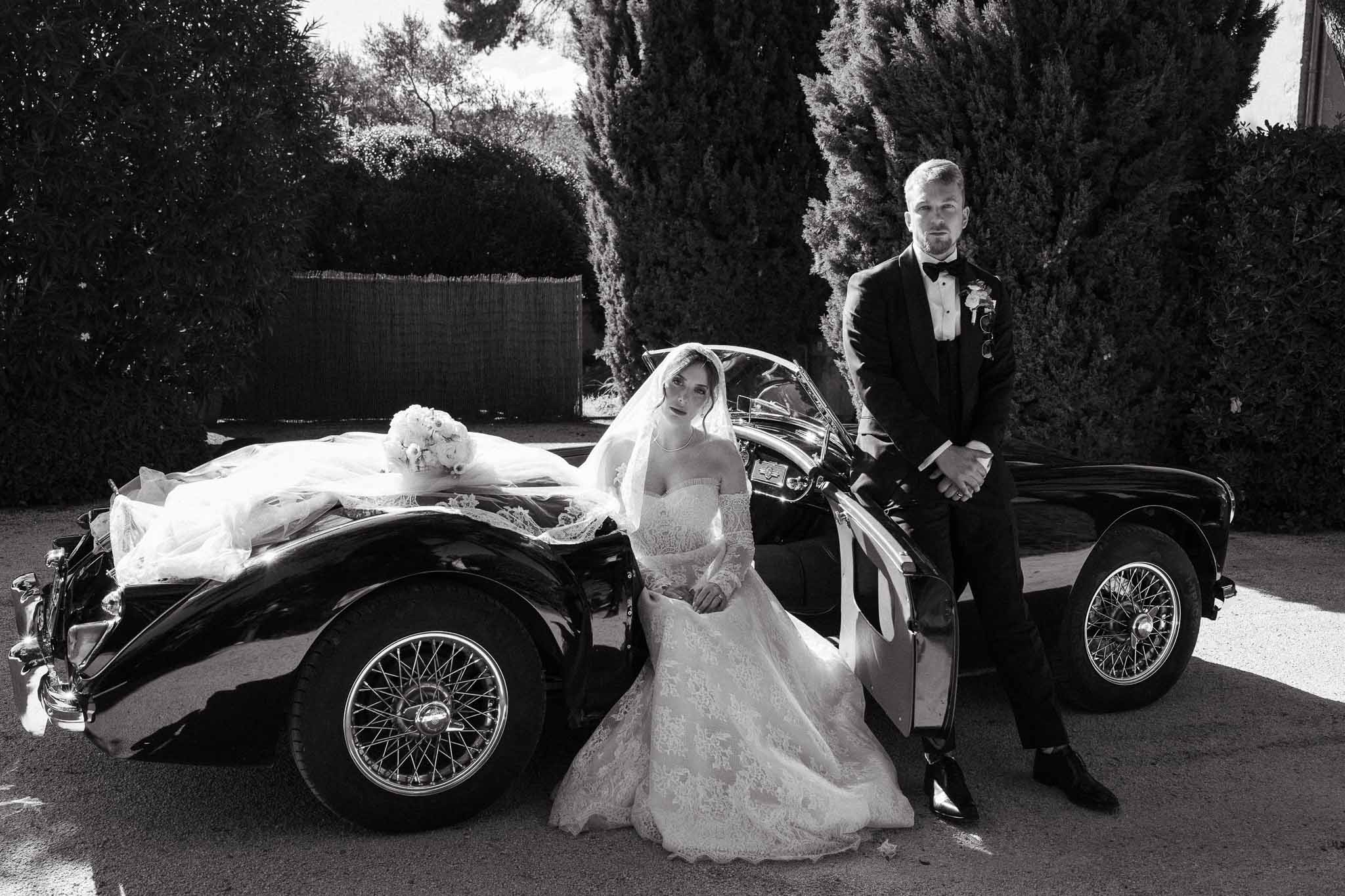 Black and white portrait of couple posed with vintage convertible sports car and cypress trees