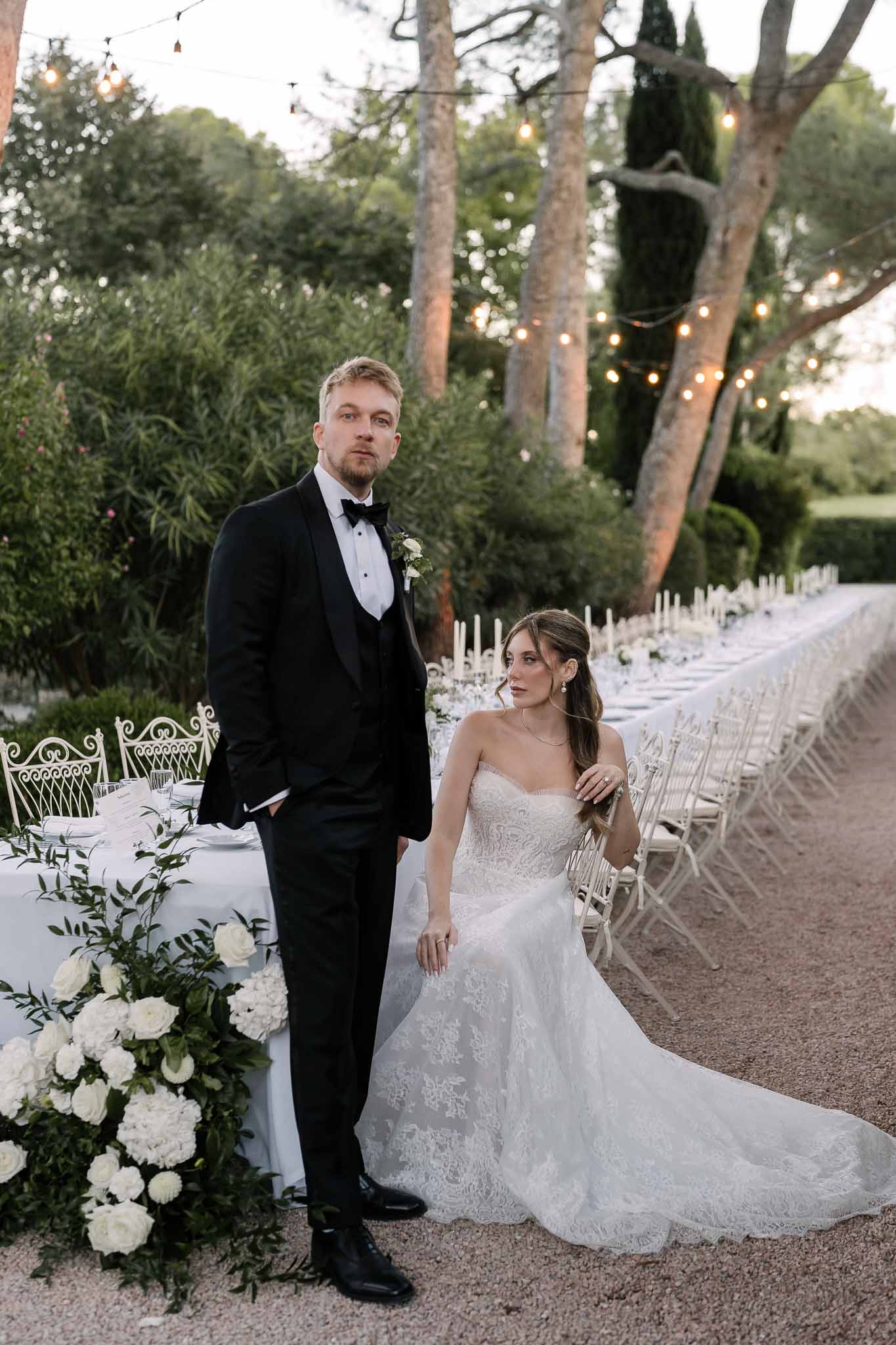 Bride seated and groom standing beside garden reception table with white candles and hydrangeas at dusk