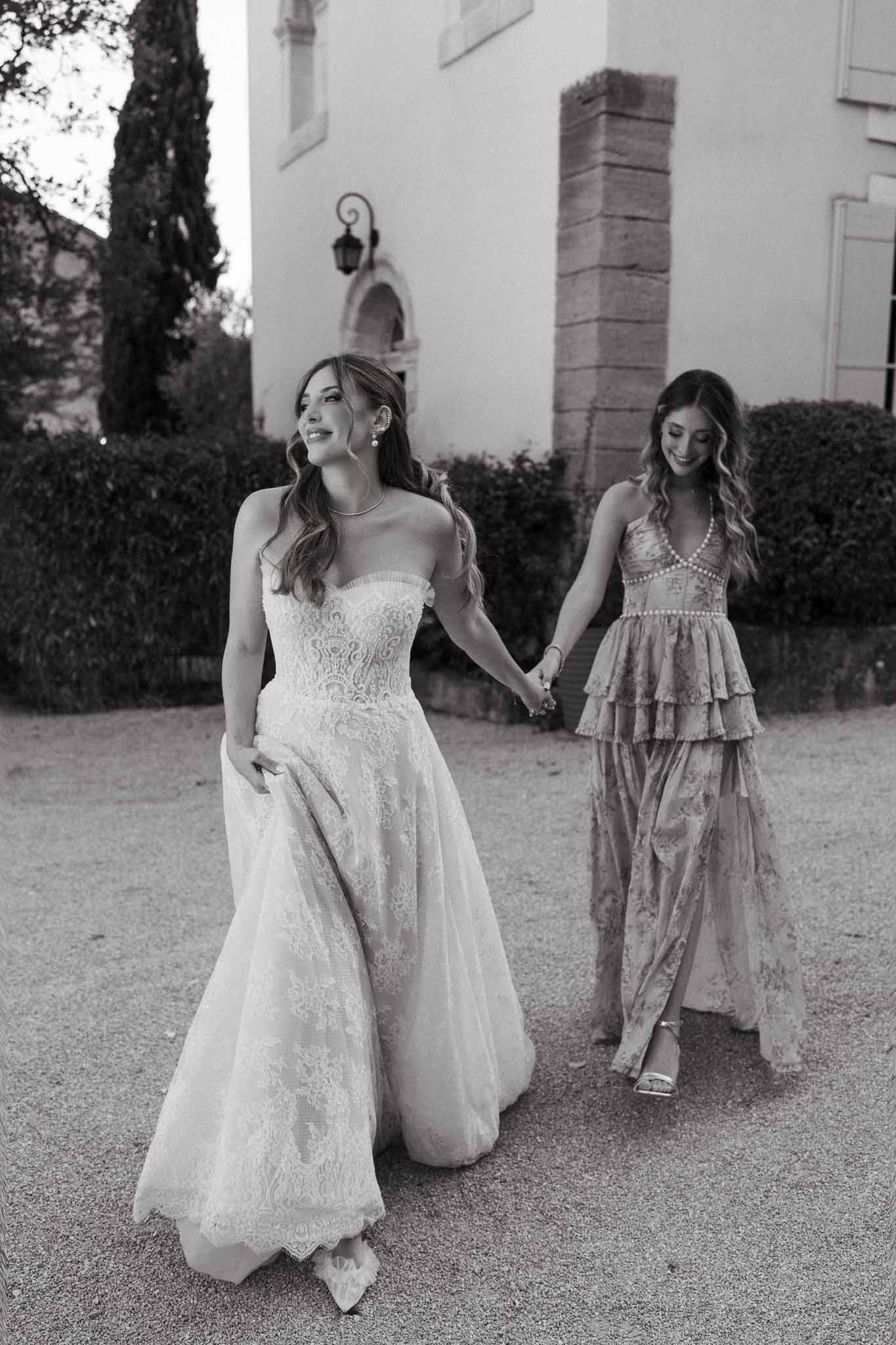 Black and white bride in strapless lace gown walking hand in hand with bridesmaid on gravel courtyard