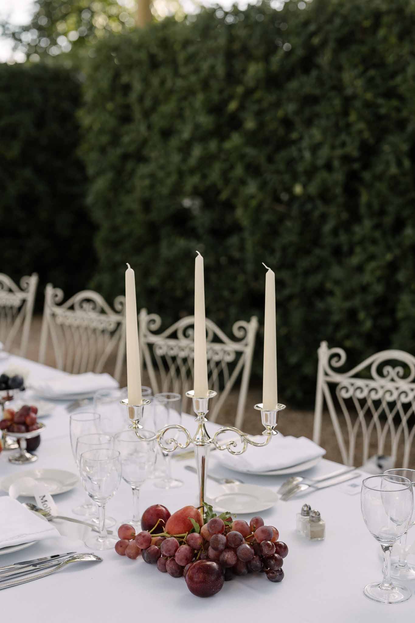 Silver three-arm candelabra with ivory tapers, red grapes, and stone fruit on white garden table