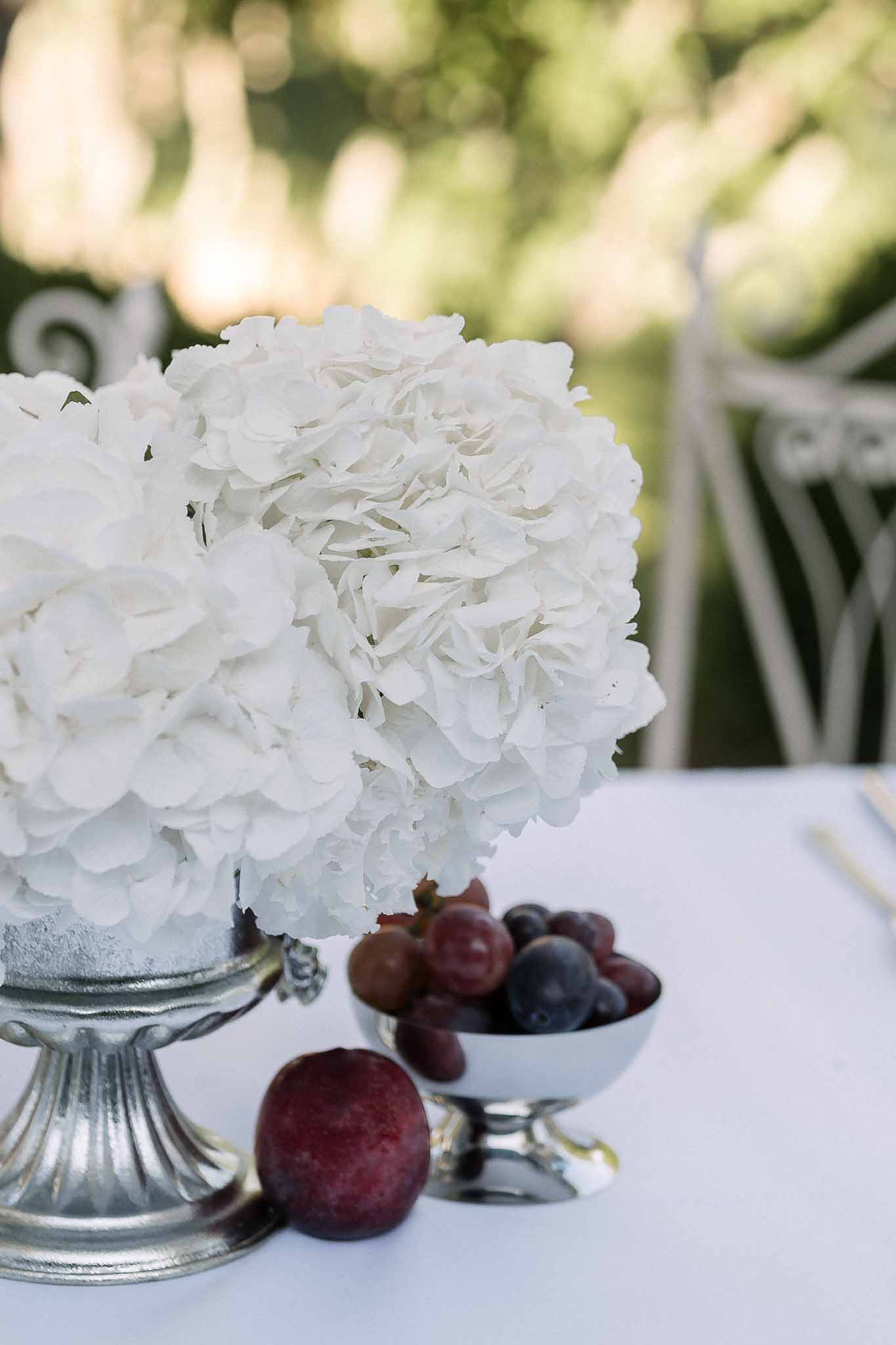 White hydrangea arrangement in silver pedestal urn beside silver bowl of dark grapes and plums