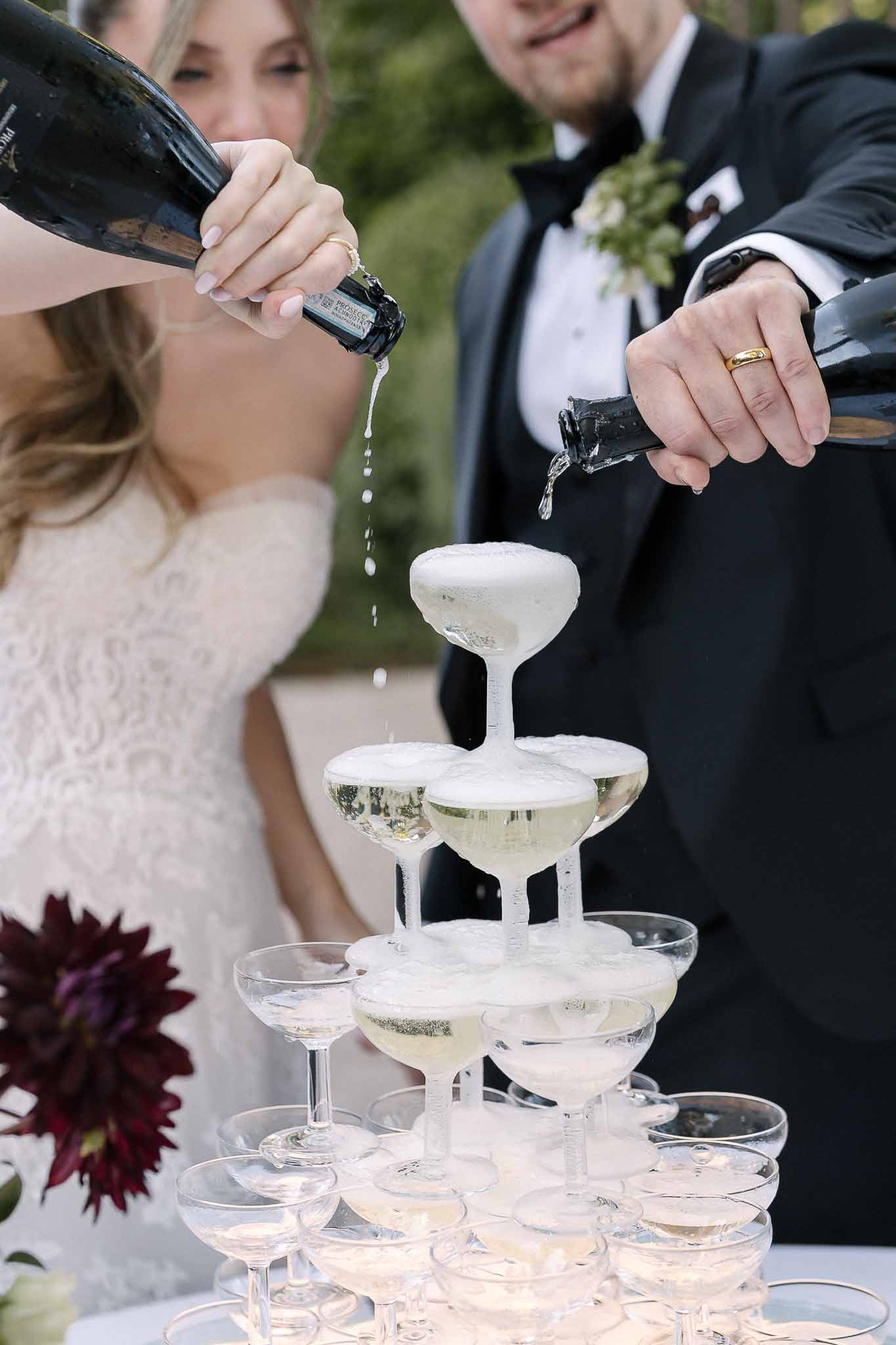 Couple pouring prosecco into cascading champagne tower with burgundy dahlia accent