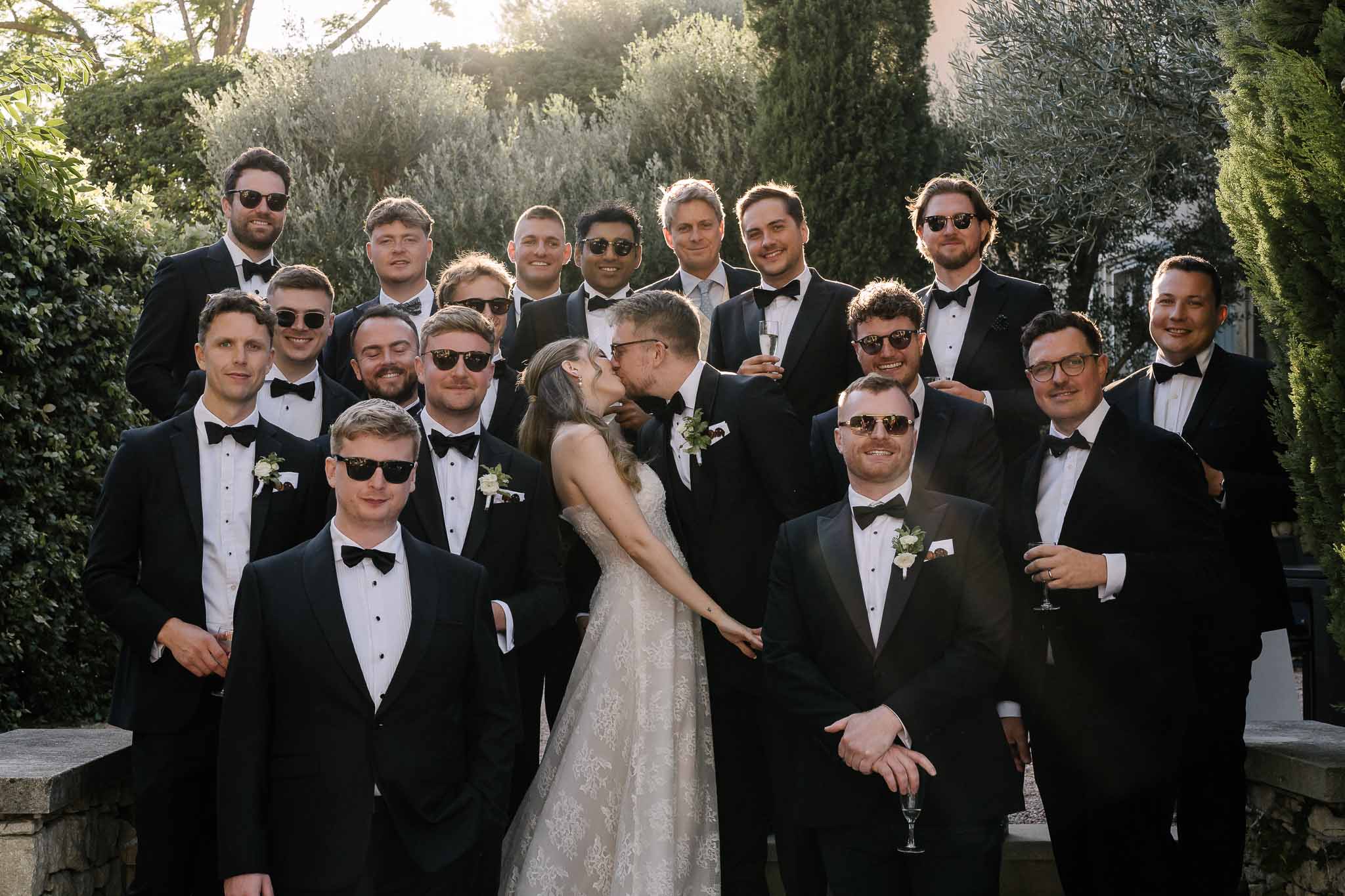Couple kissing among sixteen groomsmen in black tuxedos with sunglasses at golden hour in chateau garden