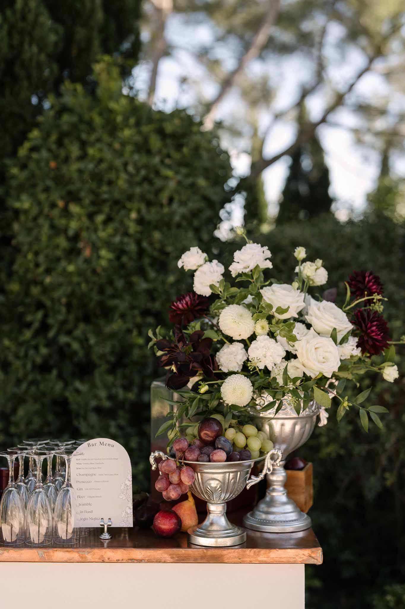 Outdoor cocktail bar with champagne flutes, printed menu, white and burgundy floral arrangement, and silver fruit bowl