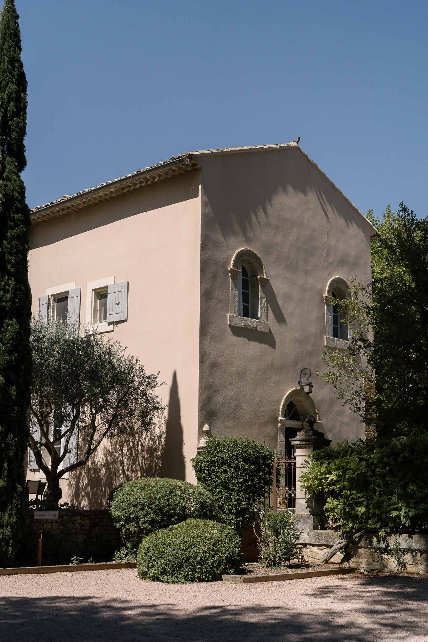 Terracotta-pink bastide with grey-blue shutters, wrought-iron gate, boxwood shrubs, and cypress trees