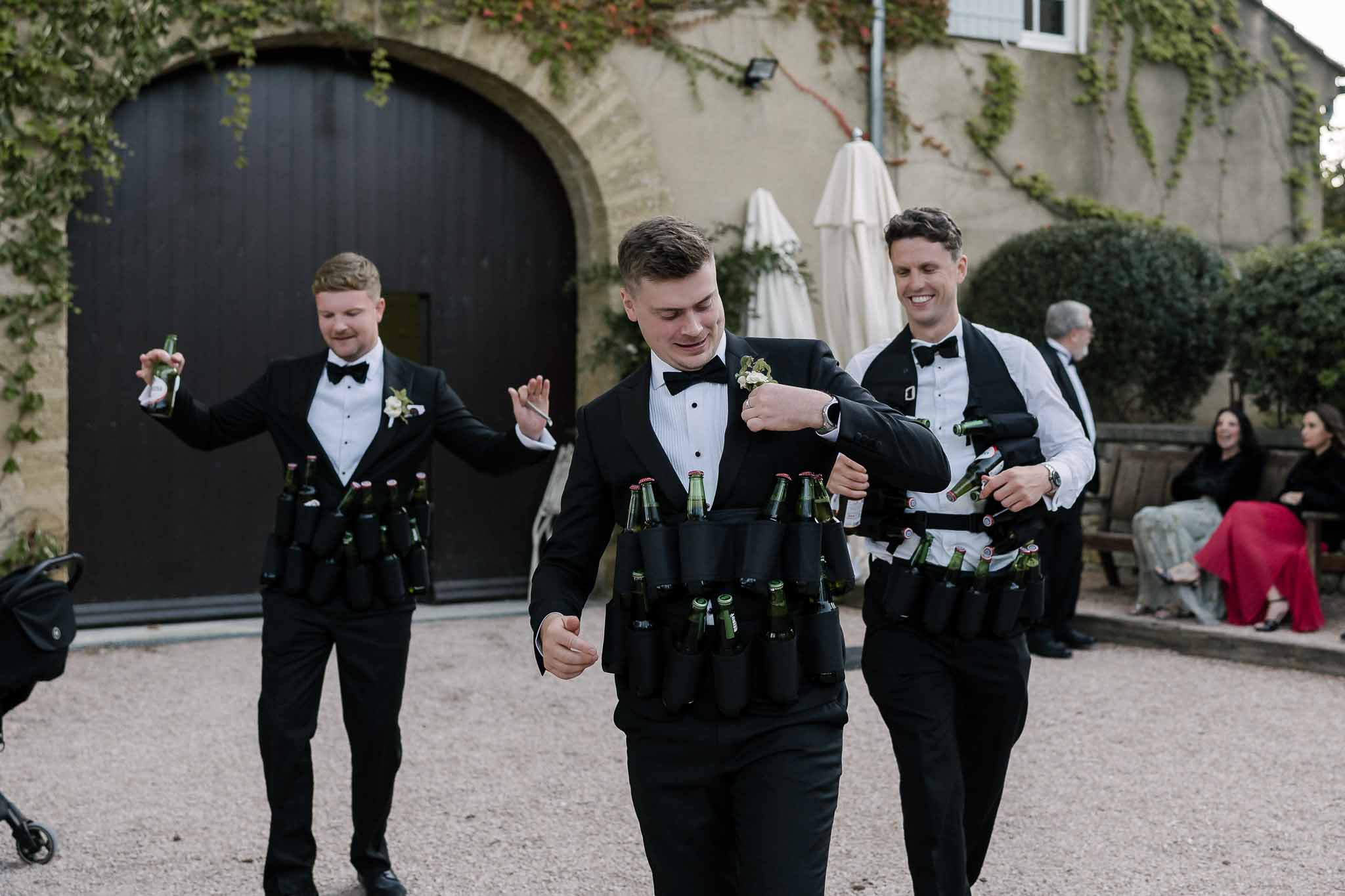 Three groomsmen in tuxedos wearing novelty beer holster vests distributing drinks in chateau courtyard
