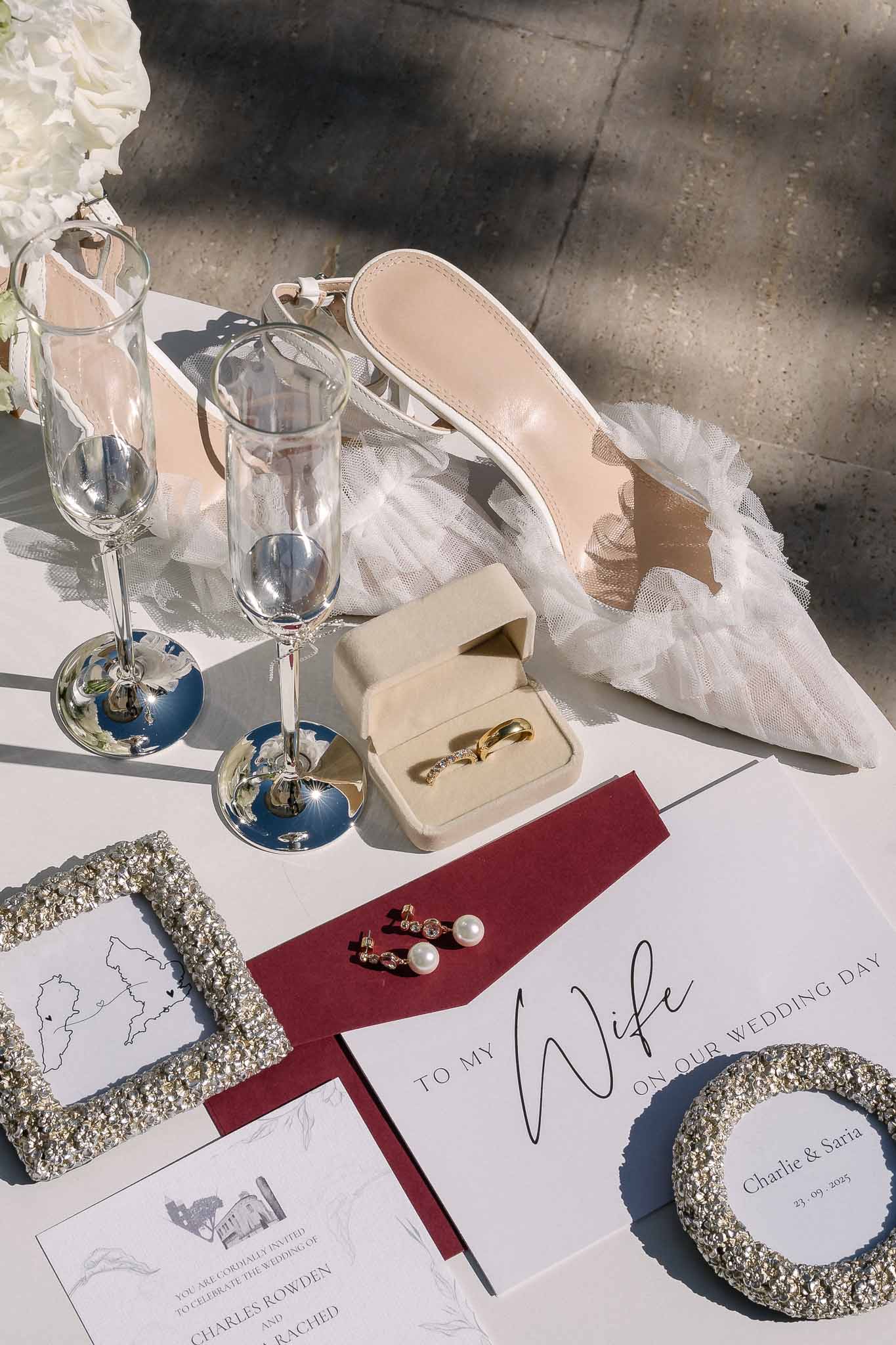 Flat lay of blush tulle-trimmed heels, gold bands in velvet box, and burgundy envelope invitation