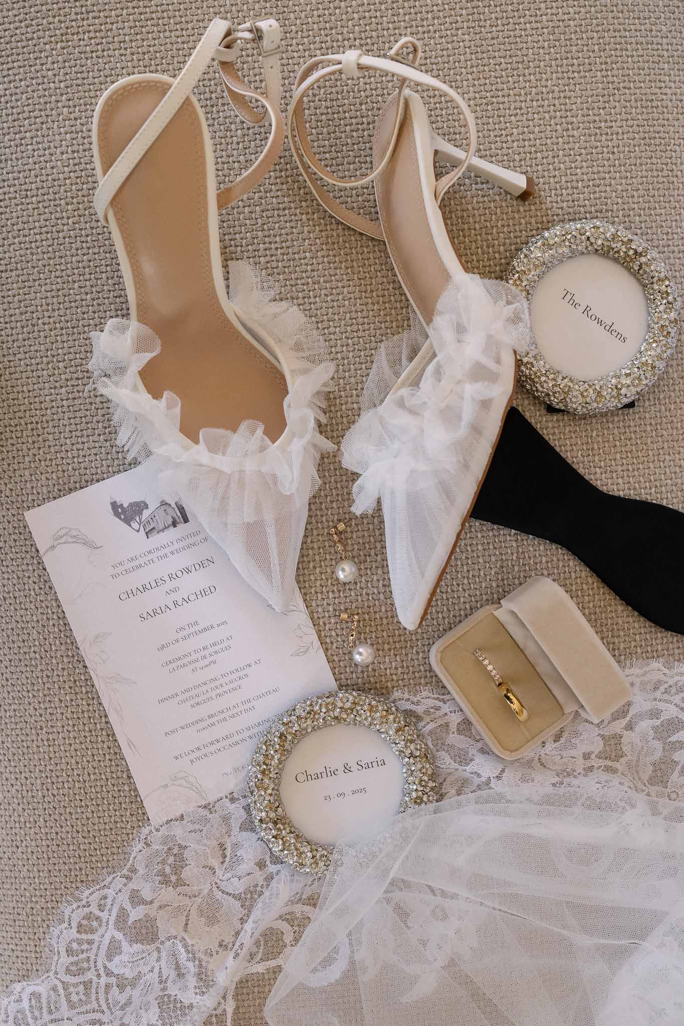 Flat lay of bridal shoes, wedding invitation, crystal frames, velvet ring box, and gold earrings on linen surface