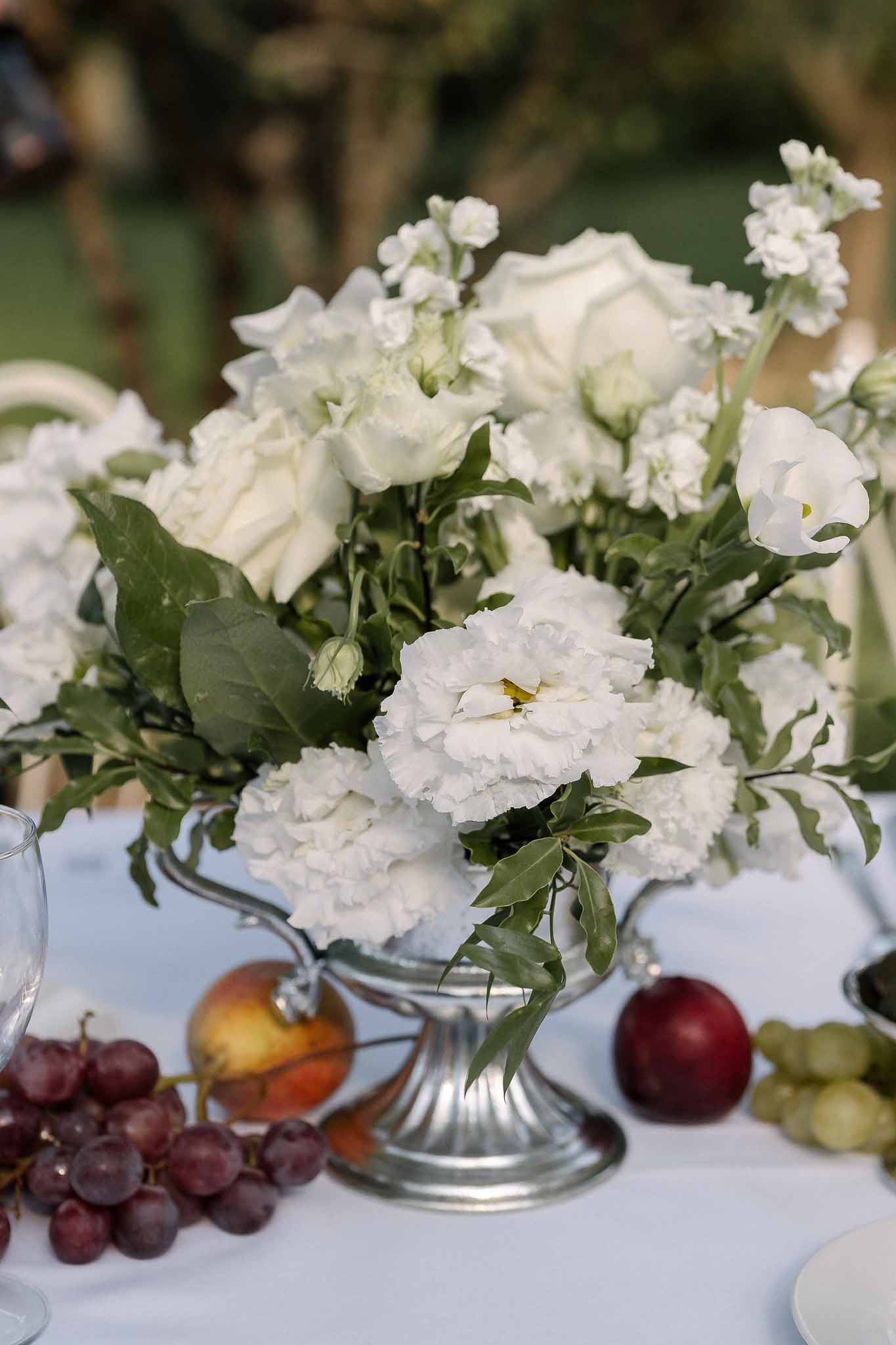 Silver urn centerpiece with white peonies, roses, and stock beside grapes and fruit on blue linen