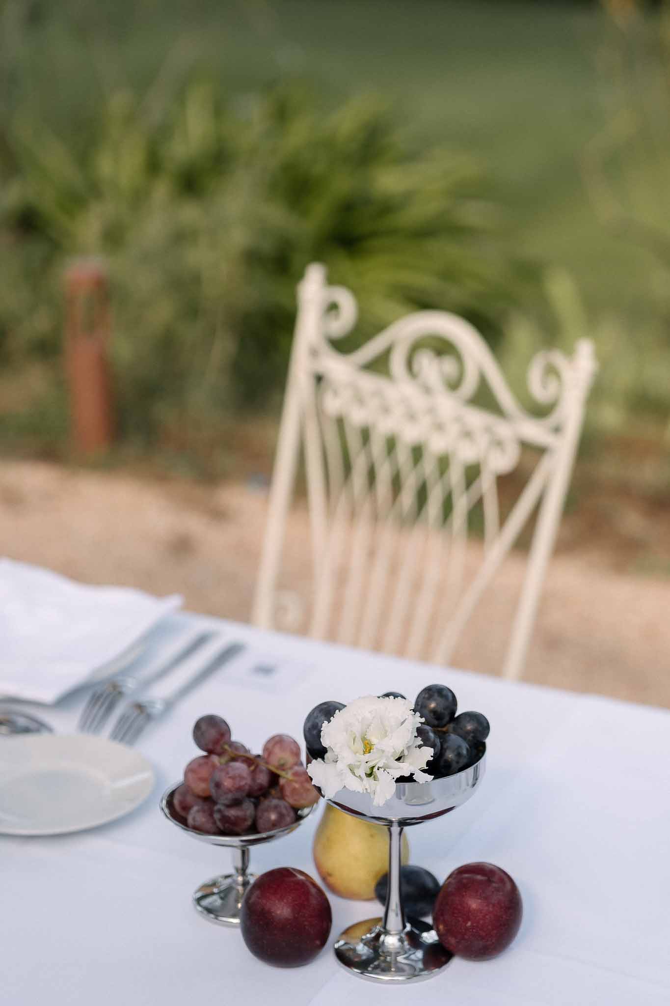 Silver coupe dishes with red and purple grapes, plums, and pear on white linen reception table